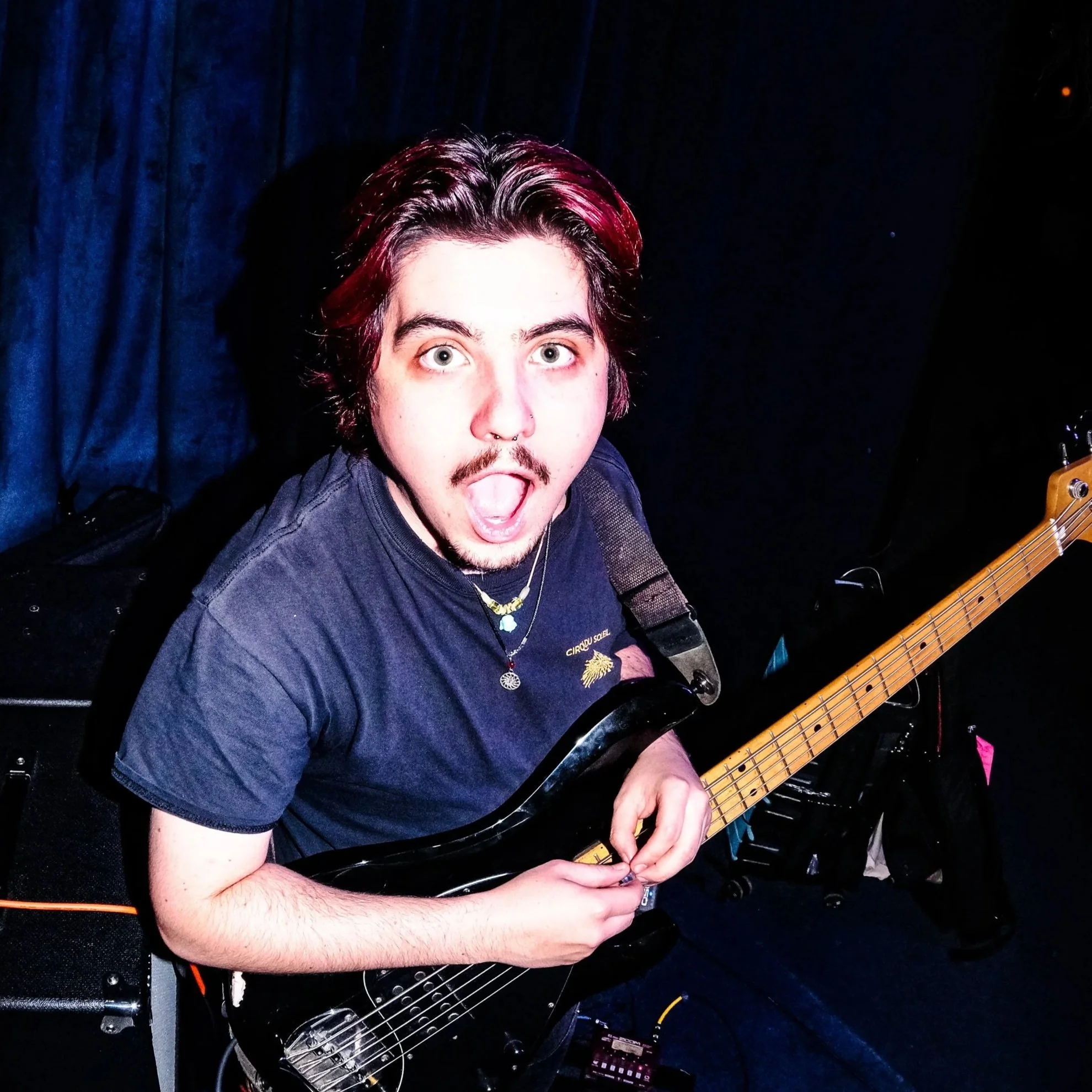 A young man with black hair, facial hair, multiple earrings, and a ring on his finger, wearing a black shirt with a subtle pattern, posing against a light gray background.