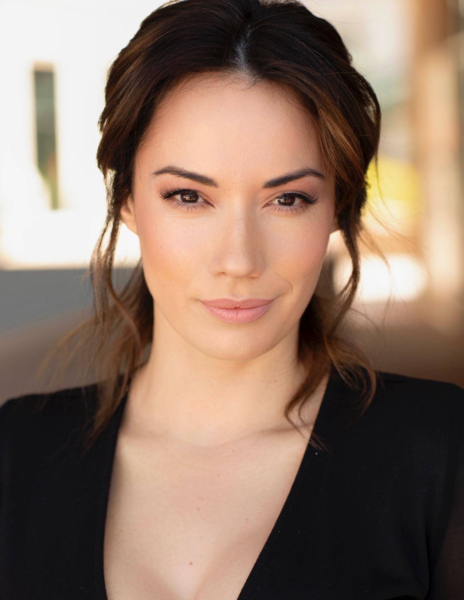 Portrait of a young woman with long, dark brown hair, fair skin, and makeup, smiling softly, against a plain gray background.
