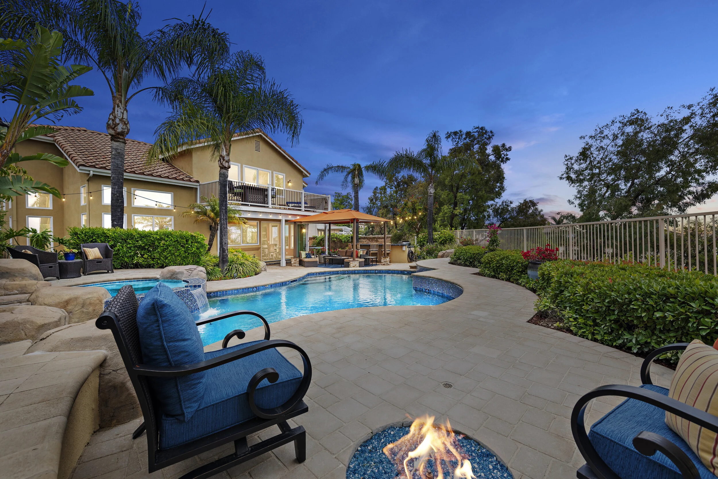 Backyard with swimming pool, patio furniture, fire pit, and house with balcony at dusk.