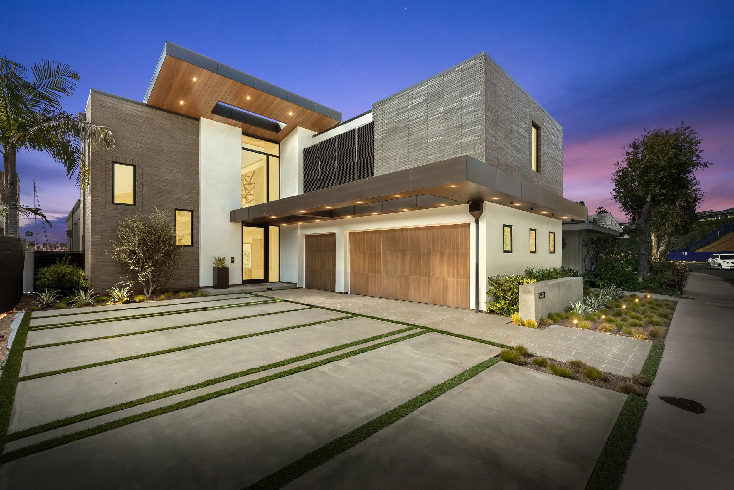 Modern house with geometric design, large windows, wooden accents, landscaped front yard, and a driveway at dusk with a purple and pink sky.