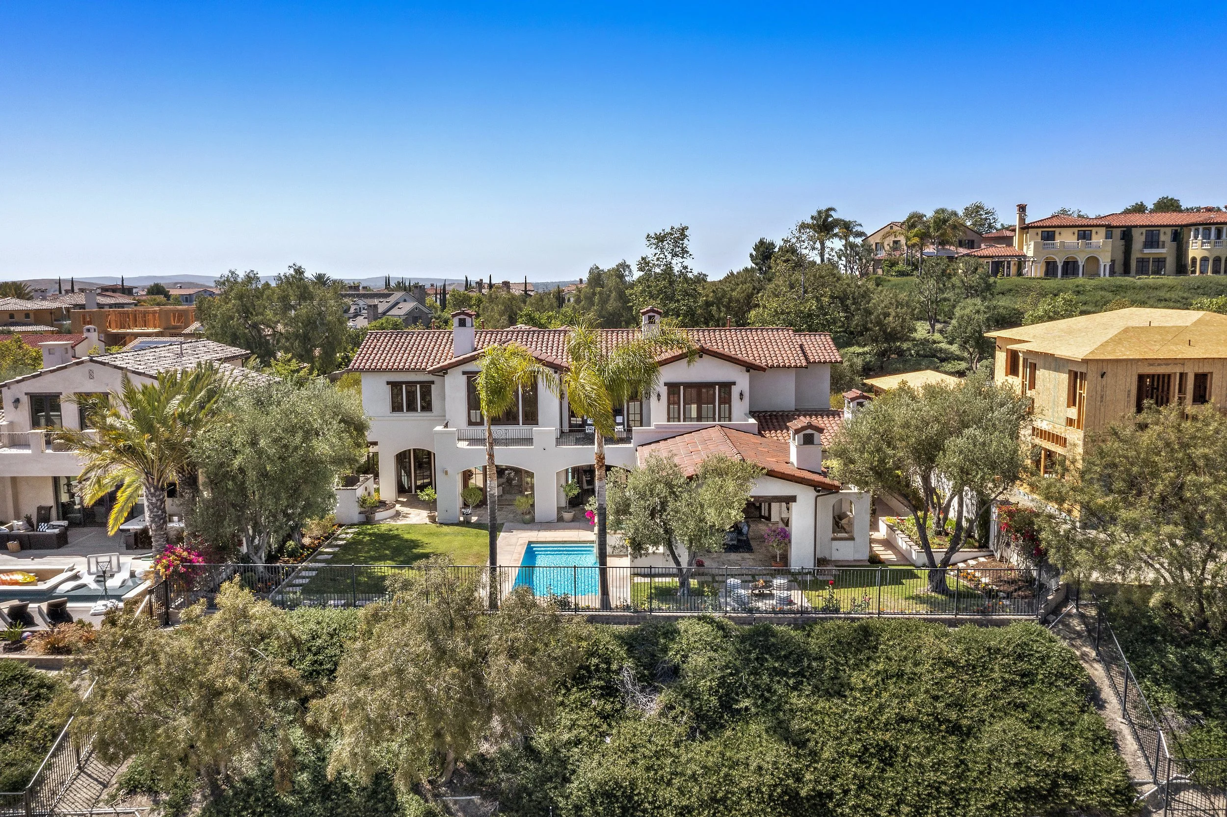 A large white house with a red tile roof and a backyard pool, surrounded by trees and other houses on a hillside under a clear blue sky.