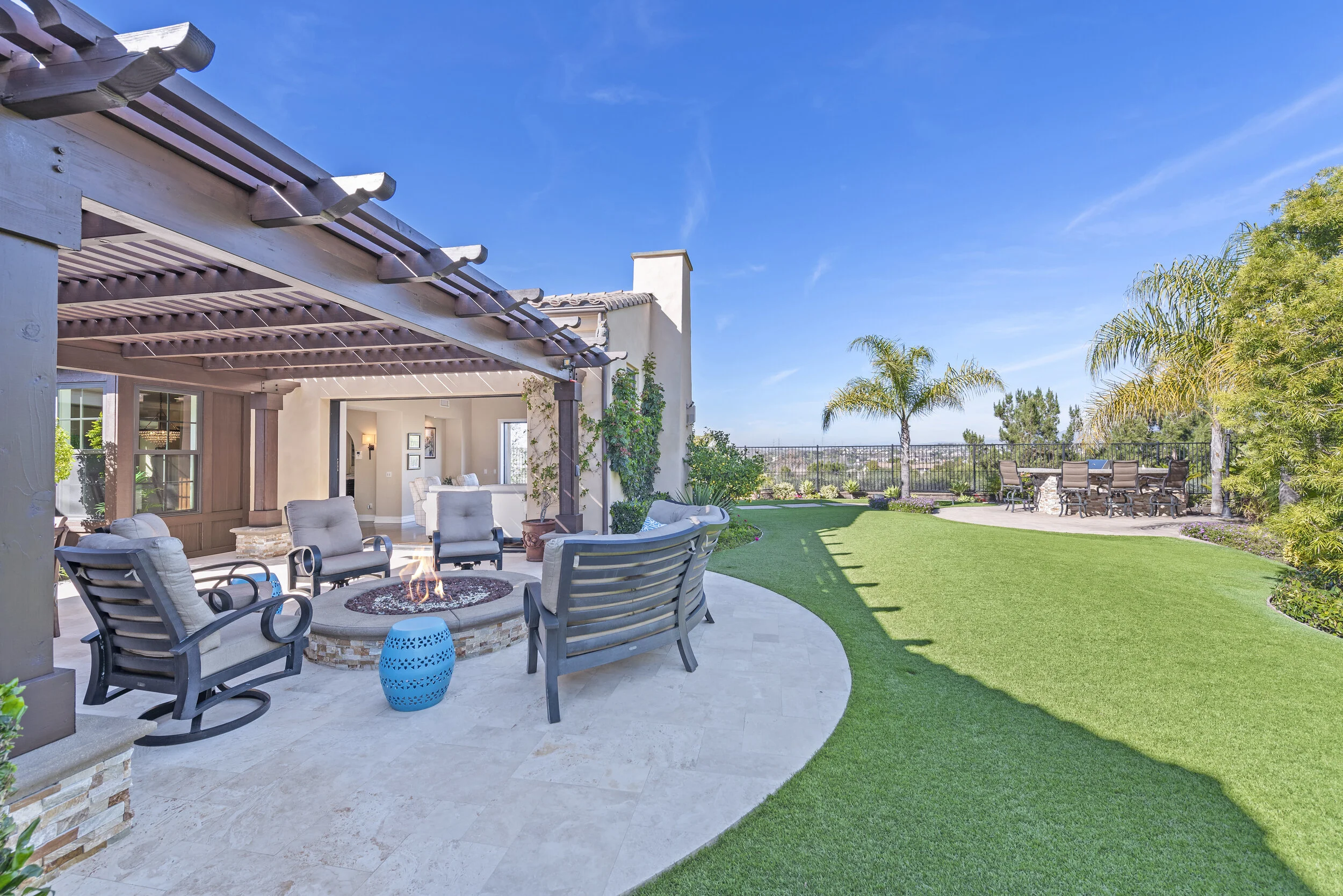 Backyard patio with a fire pit, cushioned chairs, and a covered seating area, surrounded by a lush green lawn and palm trees under a bright blue sky.