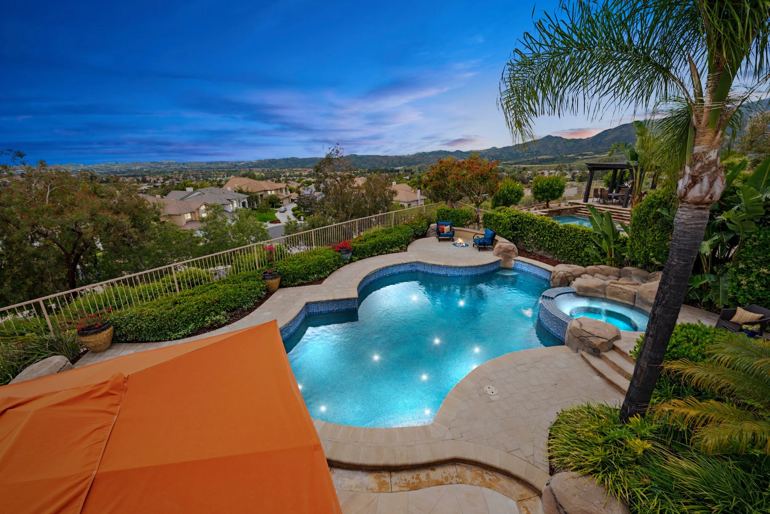 A backyard with a swimming pool, hot tub, lounge chairs, and a view of distant hills at sunset.