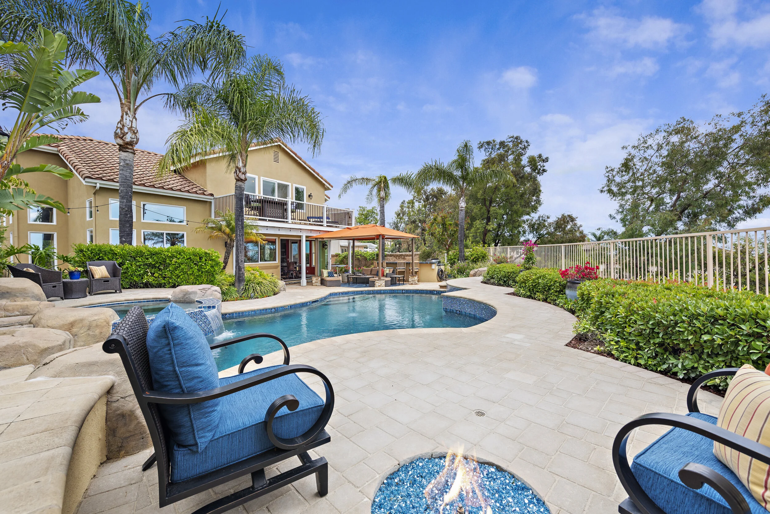 Backyard with a swimming pool, surrounded by chairs, lush greenery, palm trees, and a multi-story house with balconies and large windows. There's a fire pit with blue stones in the foreground and a shaded seating area with an orange canopy.
