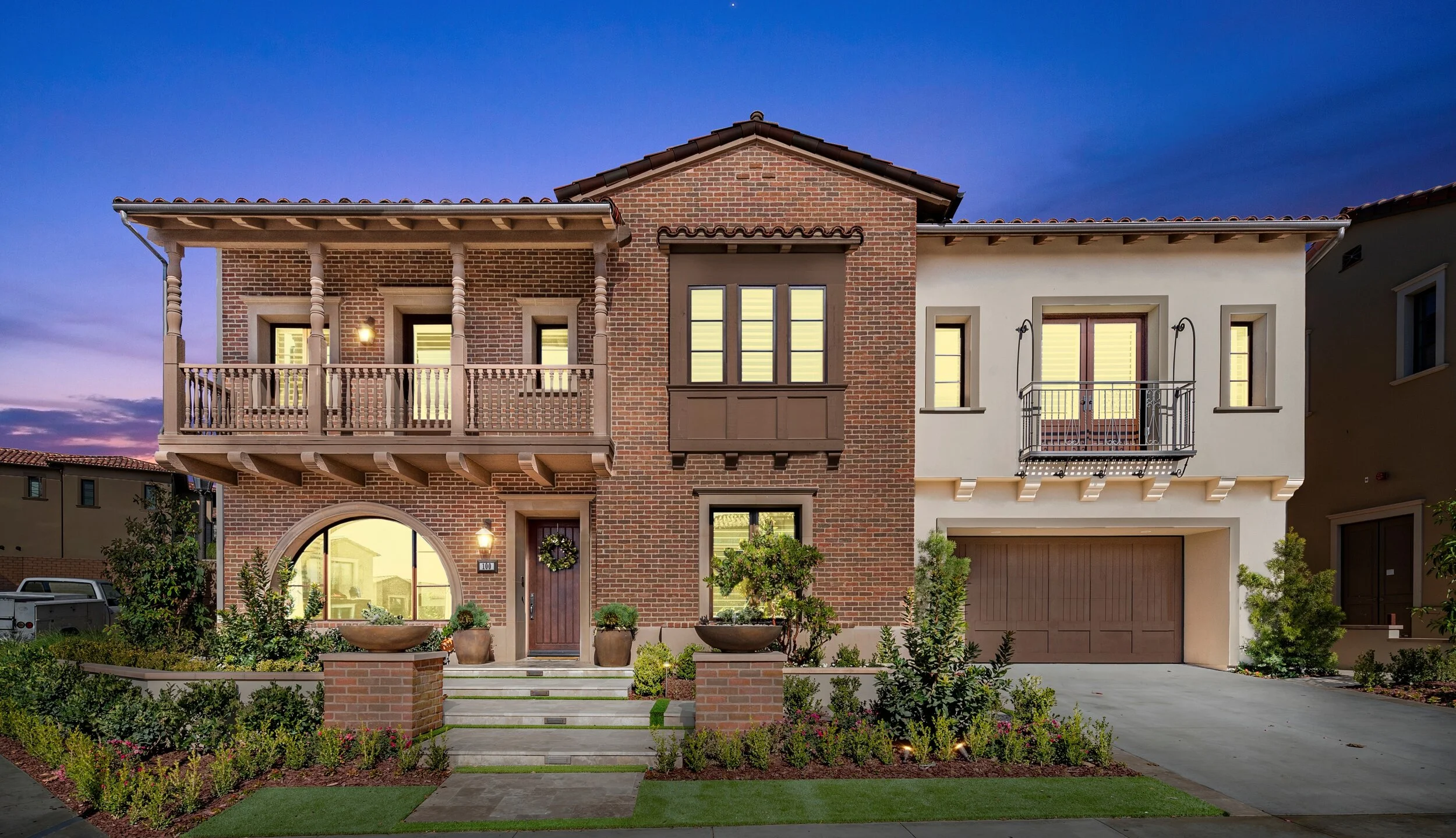 A modern two-story house with a mix of brick and stucco exterior, front porch, and landscaped garden at dusk.