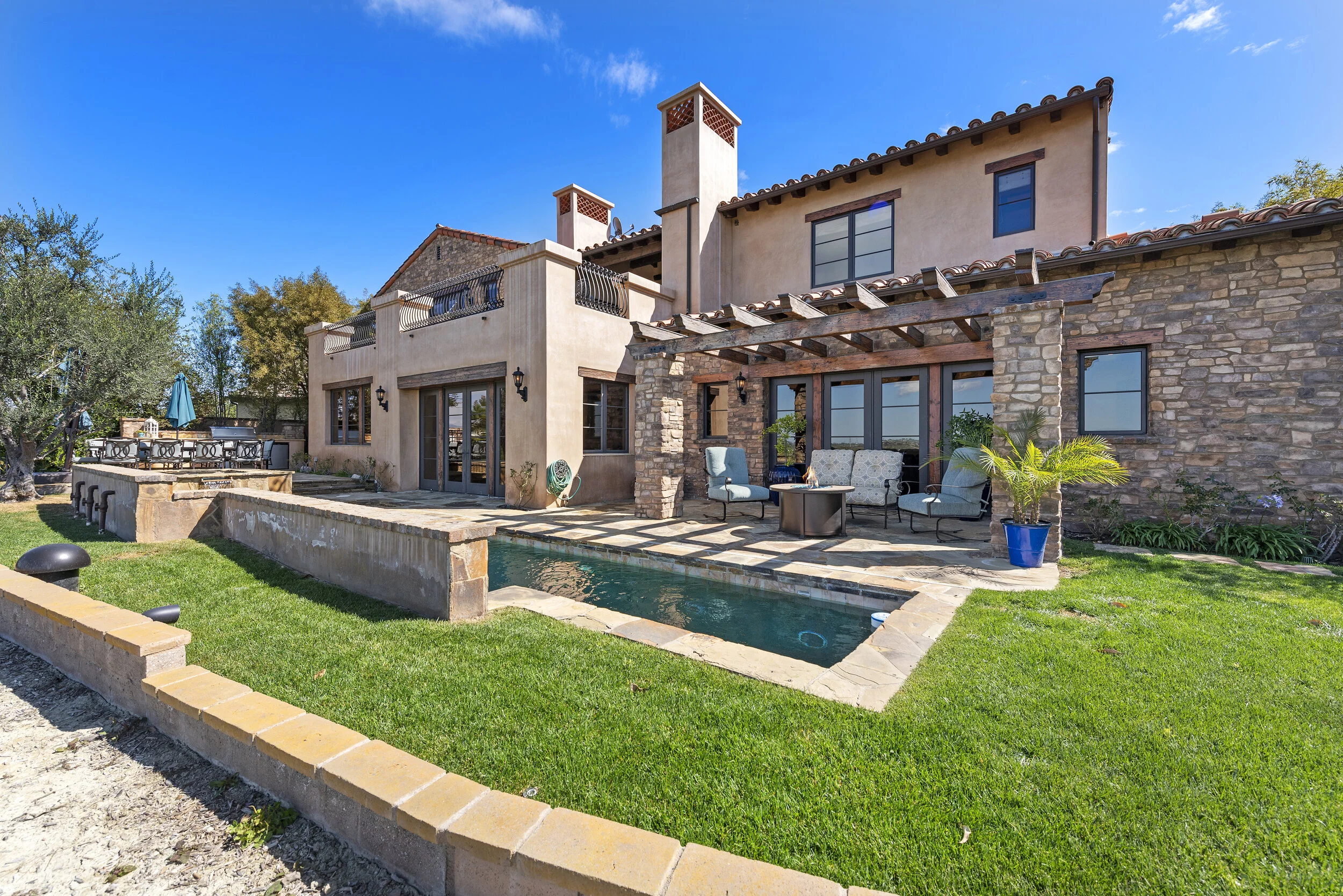 Backyard with a small pool, patio furniture, and a two-story beige and brick house under a blue sky.