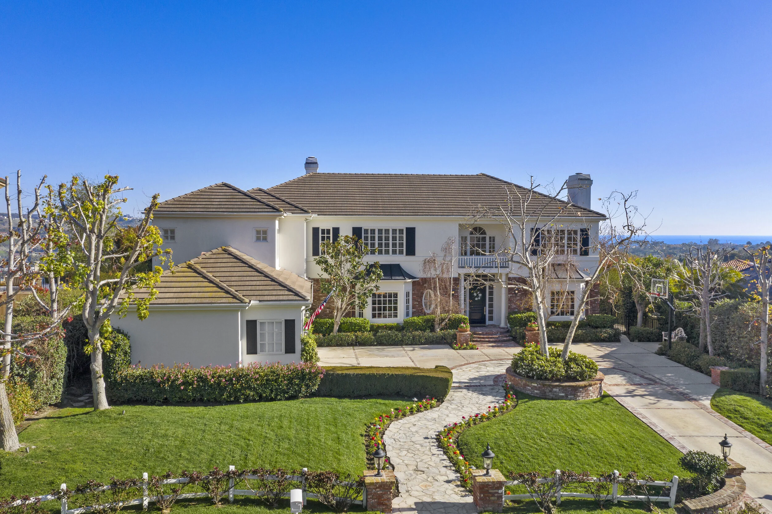 Large two-story house with white walls, brown roof, and a landscaped front yard with green grass, flowering plants, leafless trees, and a curved paved walkway leading to the front door. There are basketball hoops and distant ocean views in the backgr