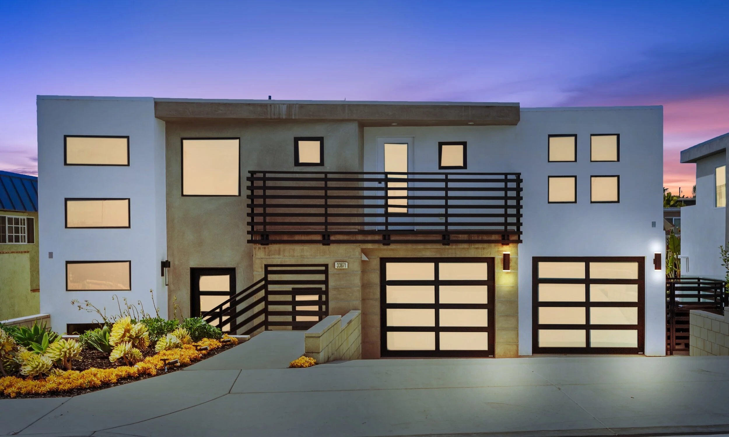 Modern multi-story house with white and beige exterior, black framed windows, wooden balcony railing, and landscaped front yard at dusk with a colorful sky.