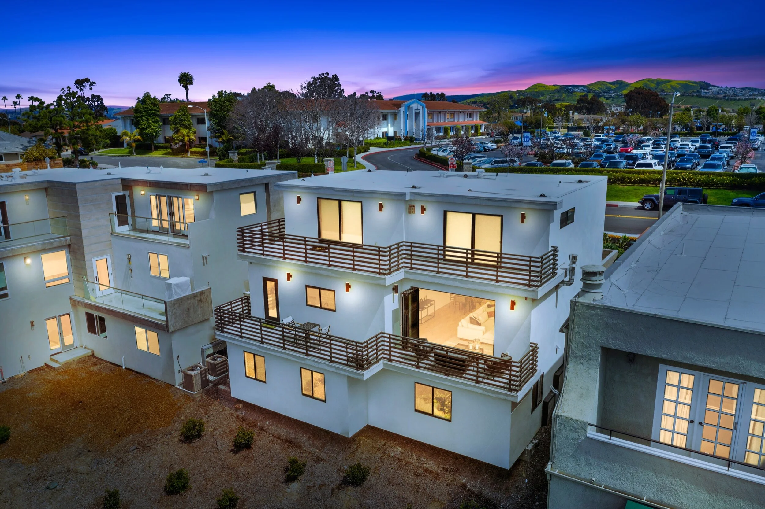 Apartment building with lit interior and balconies, sunset sky, parking lot, and green hills in the background.