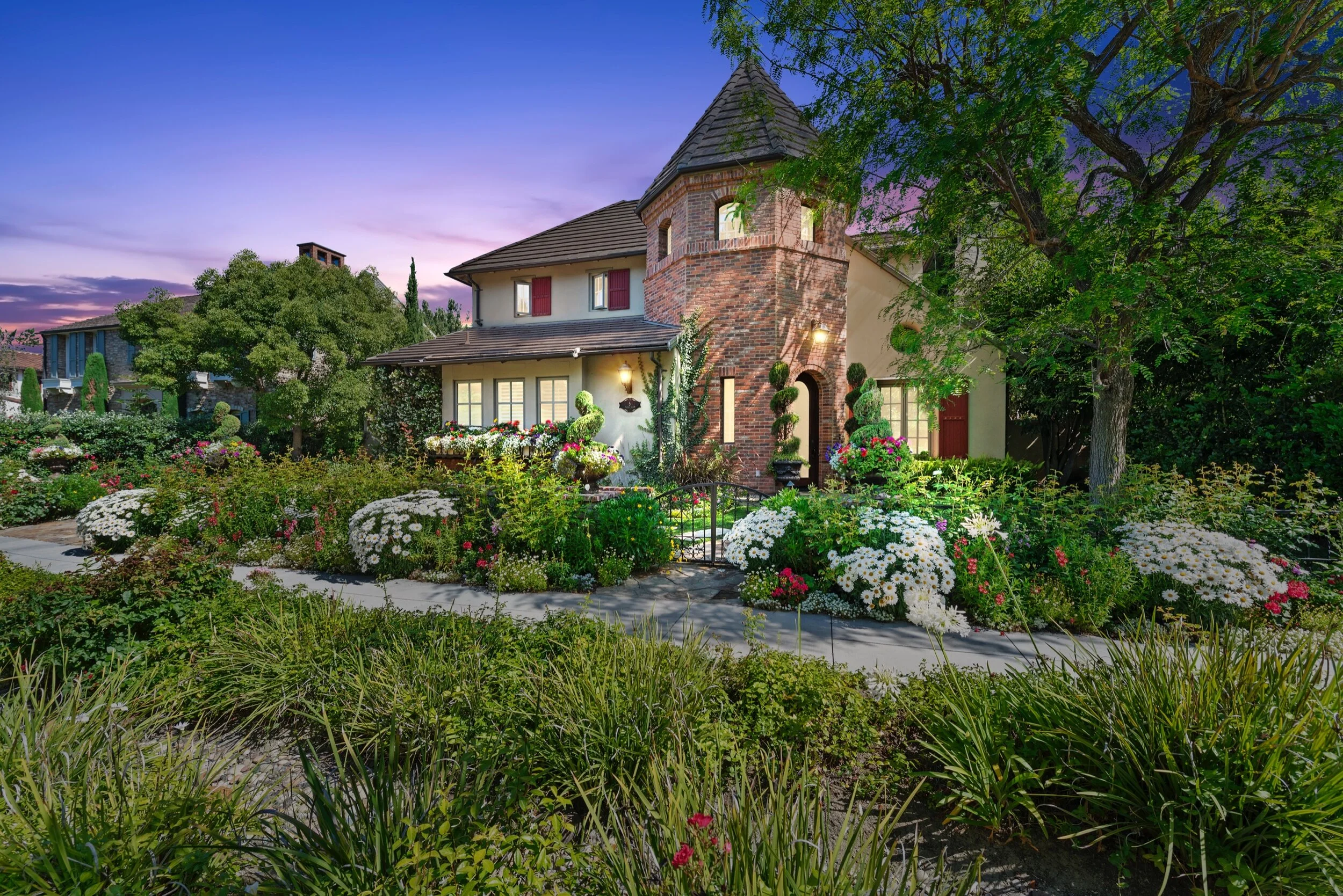 A large house with a brick turret surrounded by a well-maintained garden with colorful flowers and trees at dusk.