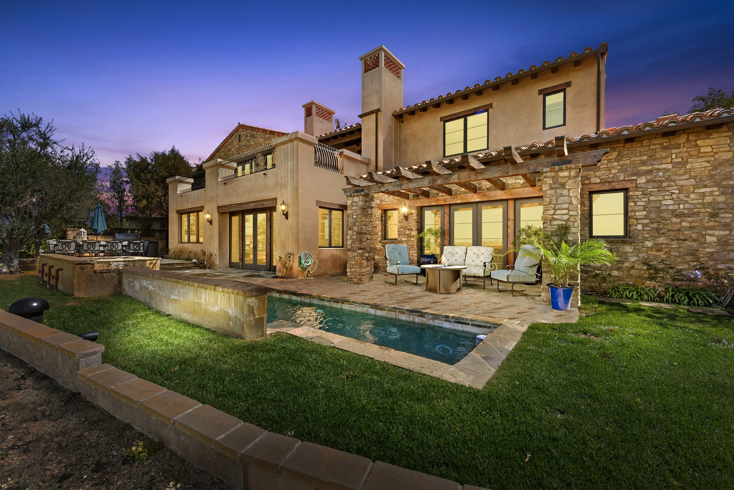 Backyard of a house with a small pool, outdoor seating area, brick and stone exterior, and a sunset sky.