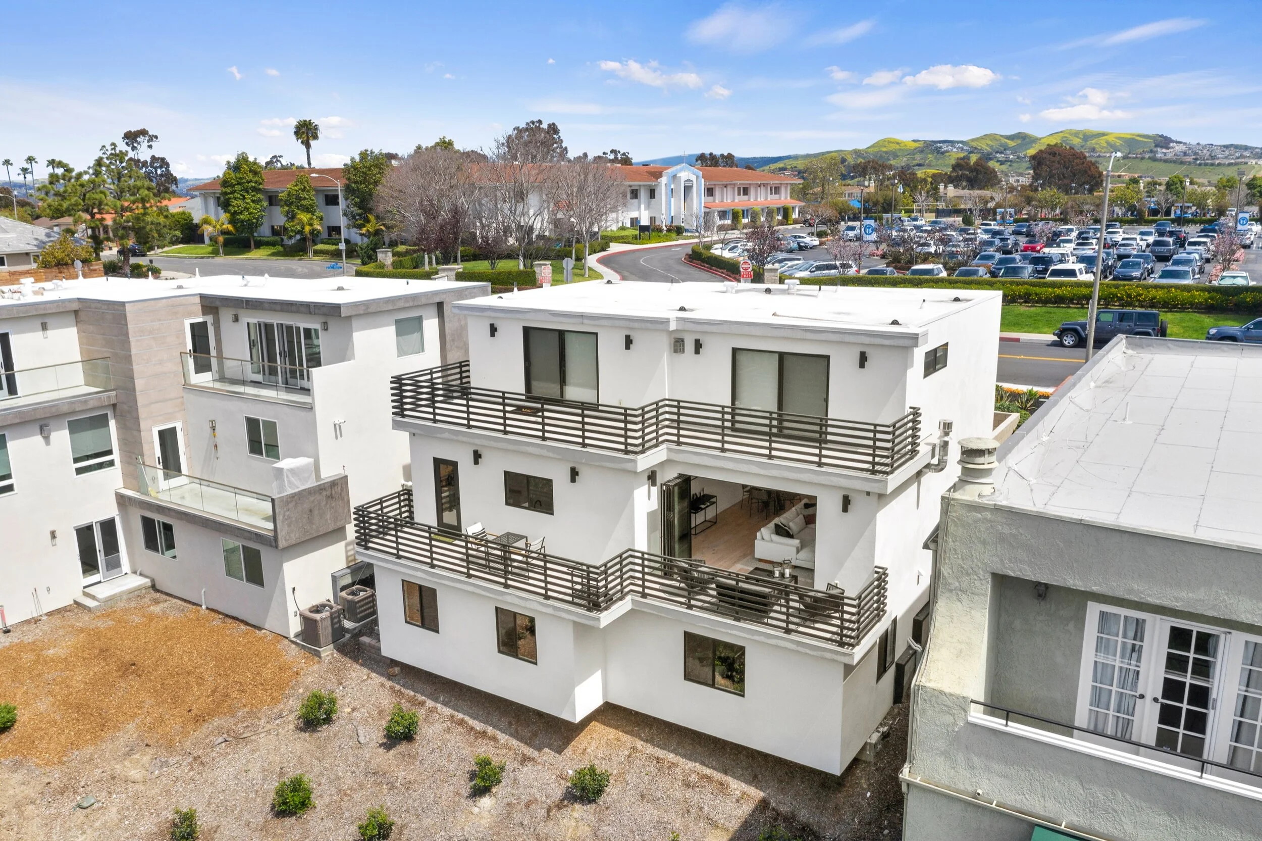Modern white multi-story apartment building with large balconies, surrounded by other residential buildings, parking lot, green hills, and blue sky with scattered clouds.