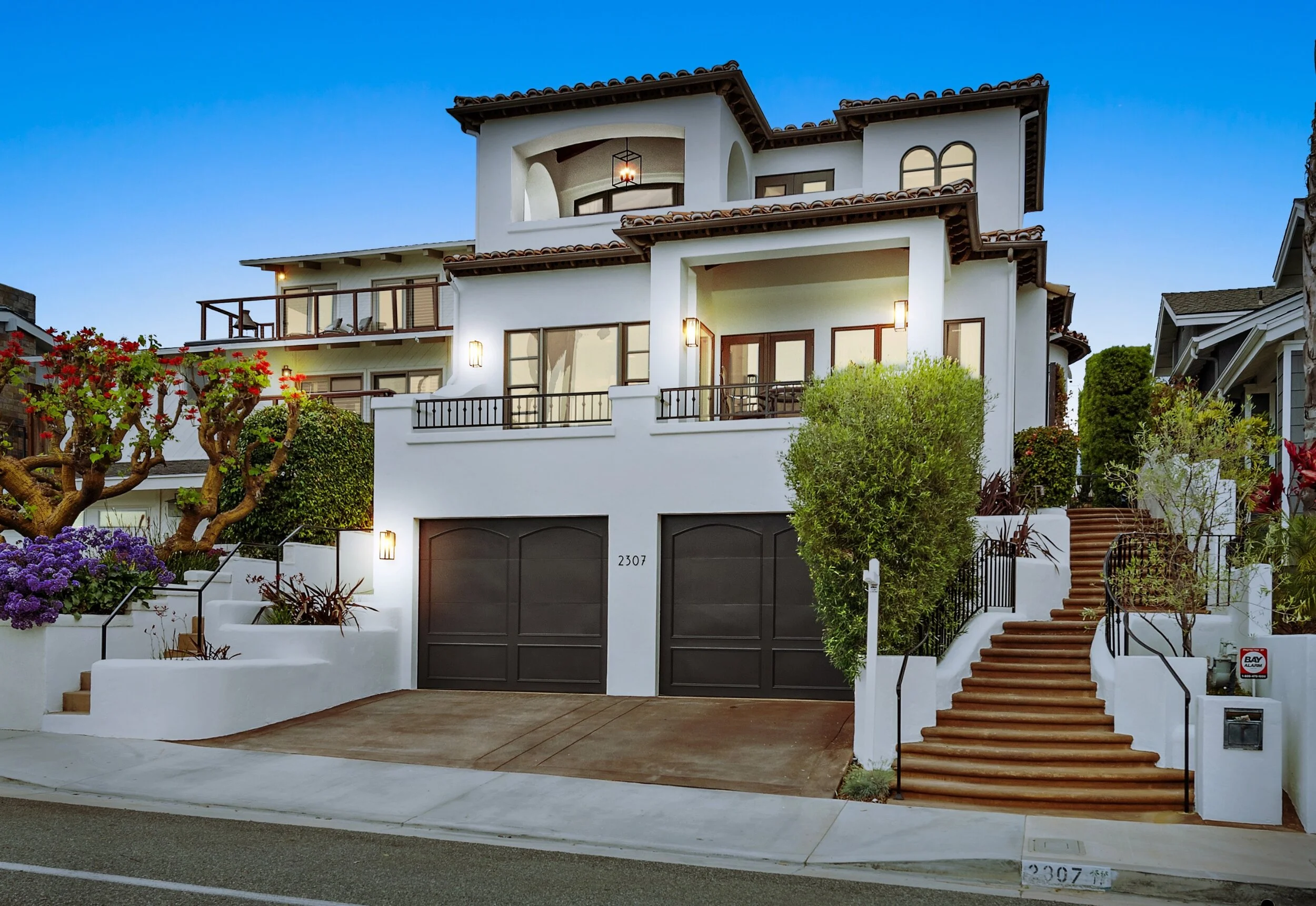 A multi-story modern house with white exterior walls, brown tile roof, balconies, and a driveway with two garage doors at street level, surrounded by landscaped plants and trees.
