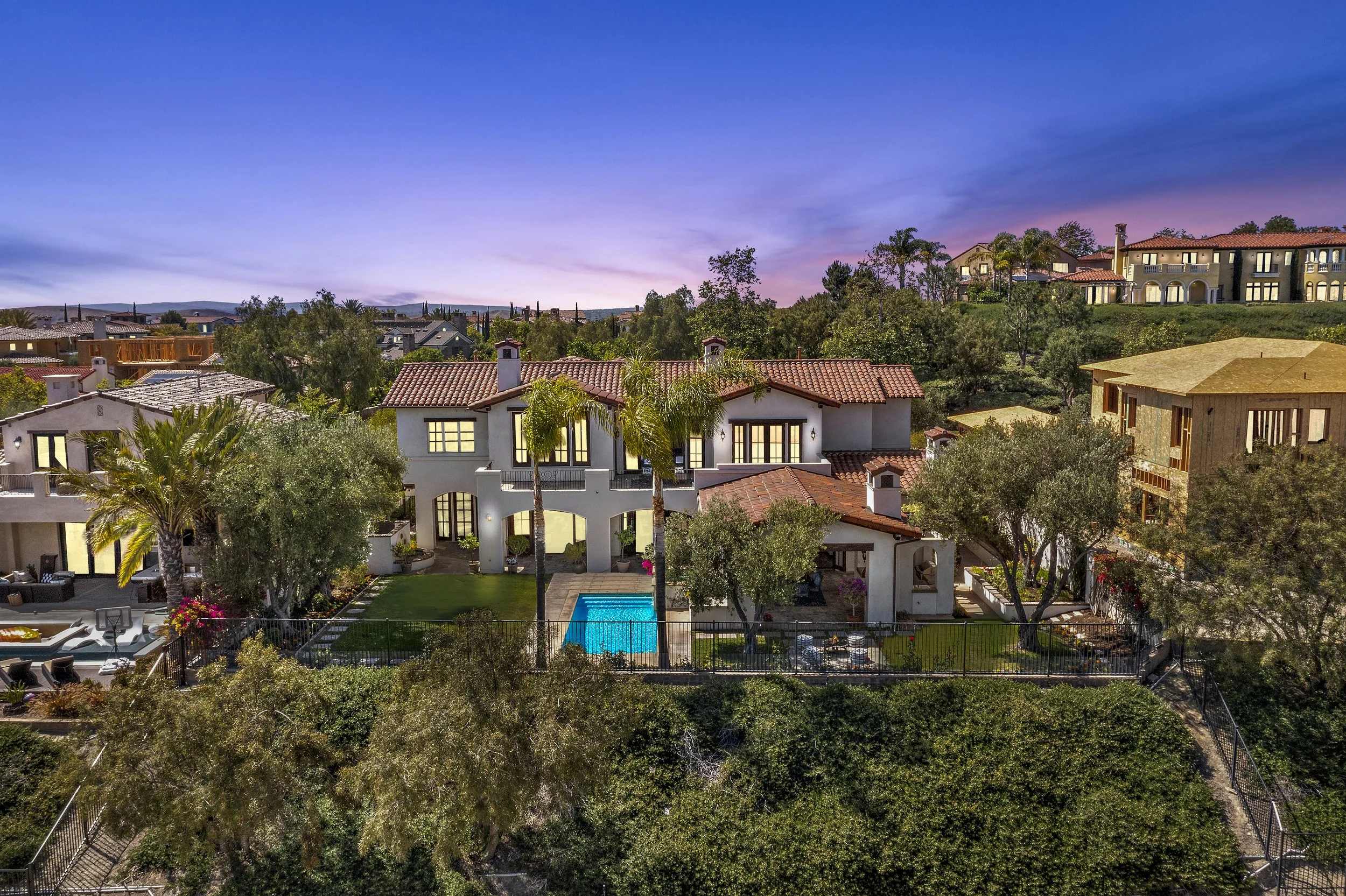 A large, luxurious house with a red tile roof, surrounded by lush trees and a fenced backyard with a small swimming pool at sunset.