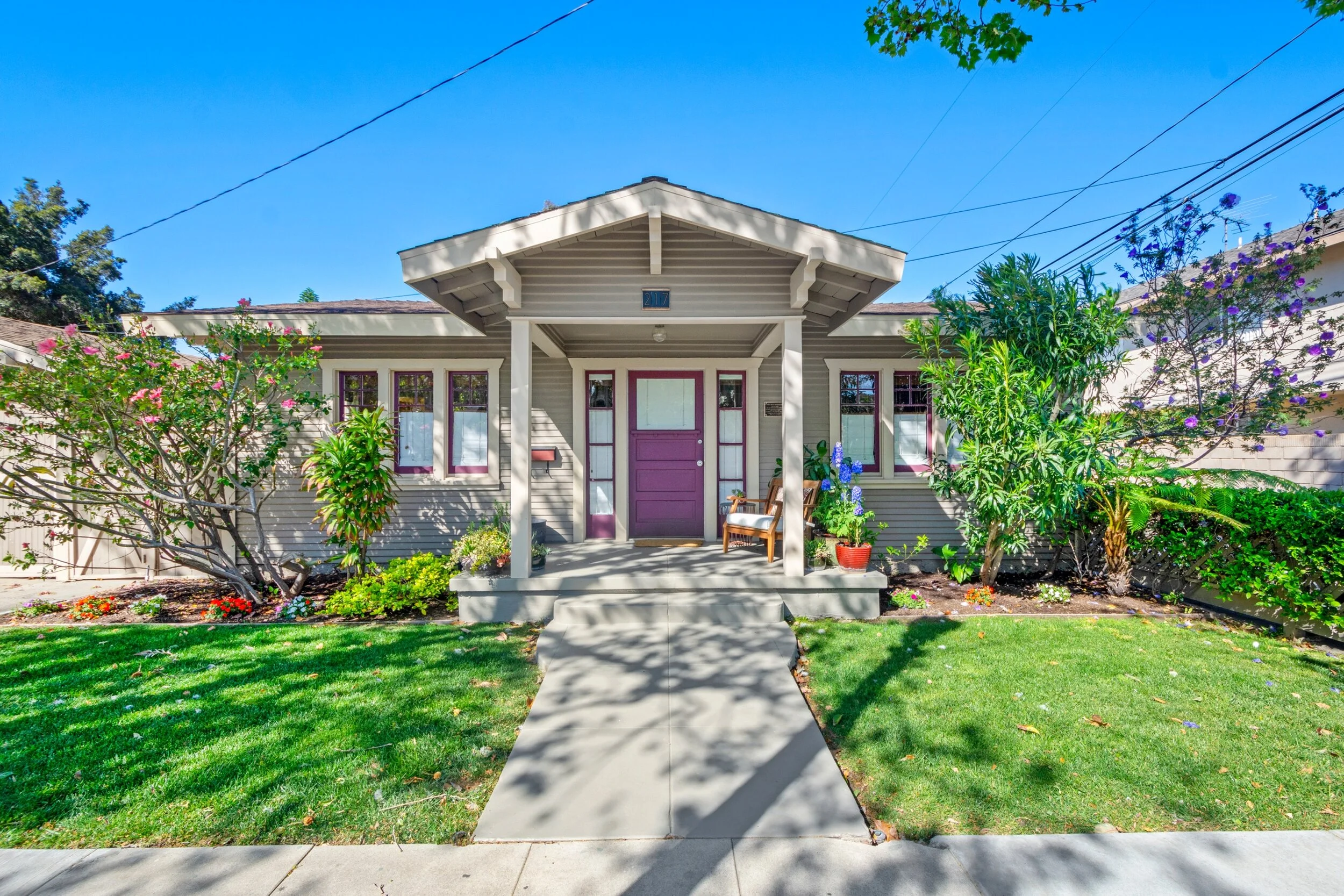 Front view of a cozy house with a purple door, front porch with two wooden chairs, well-manicured lawn, colorful flowering bushes, and a clear blue sky.