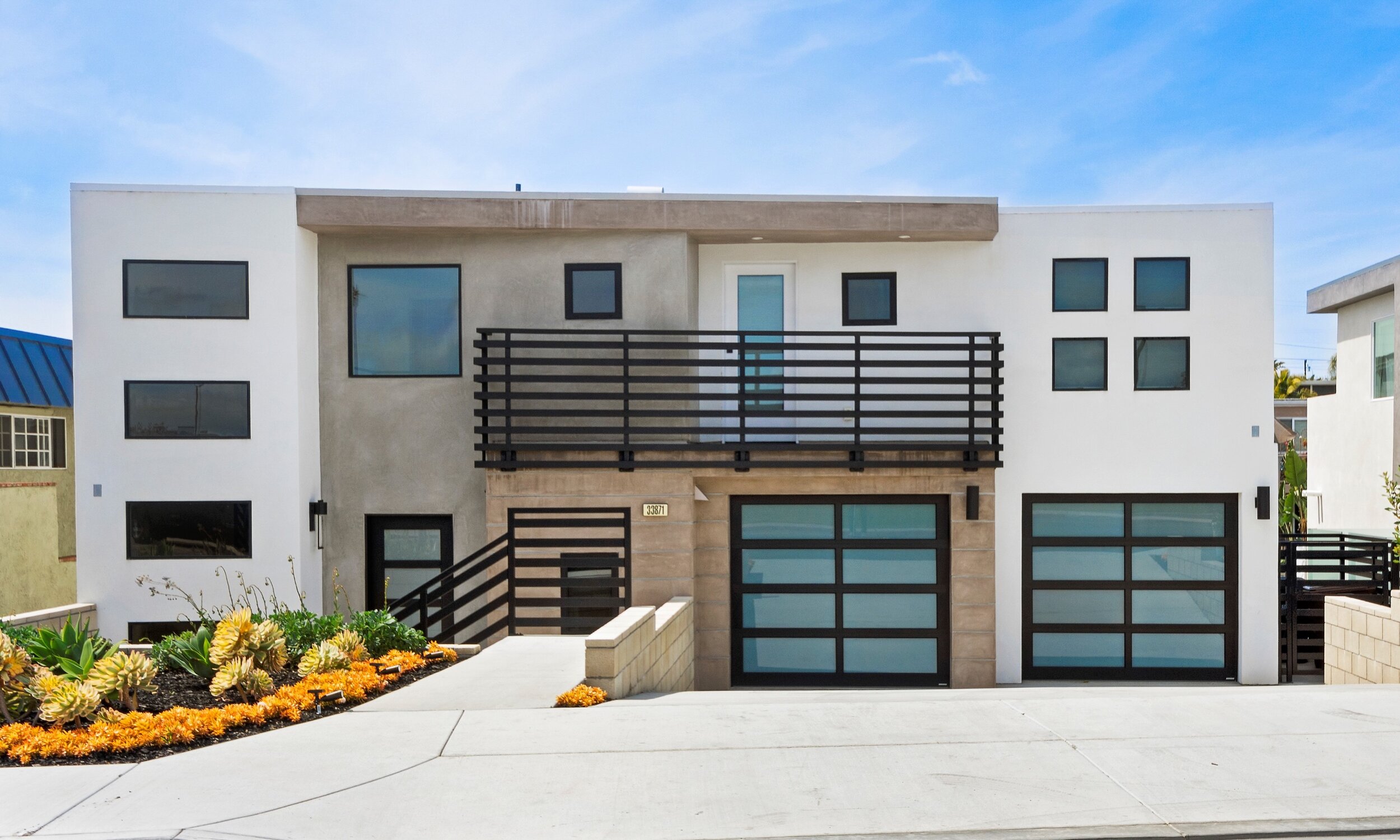 Modern multi-story house with white and gray exterior, large garage doors, black metal railings, and a flower bed with orange and yellow flowers in the front yard.