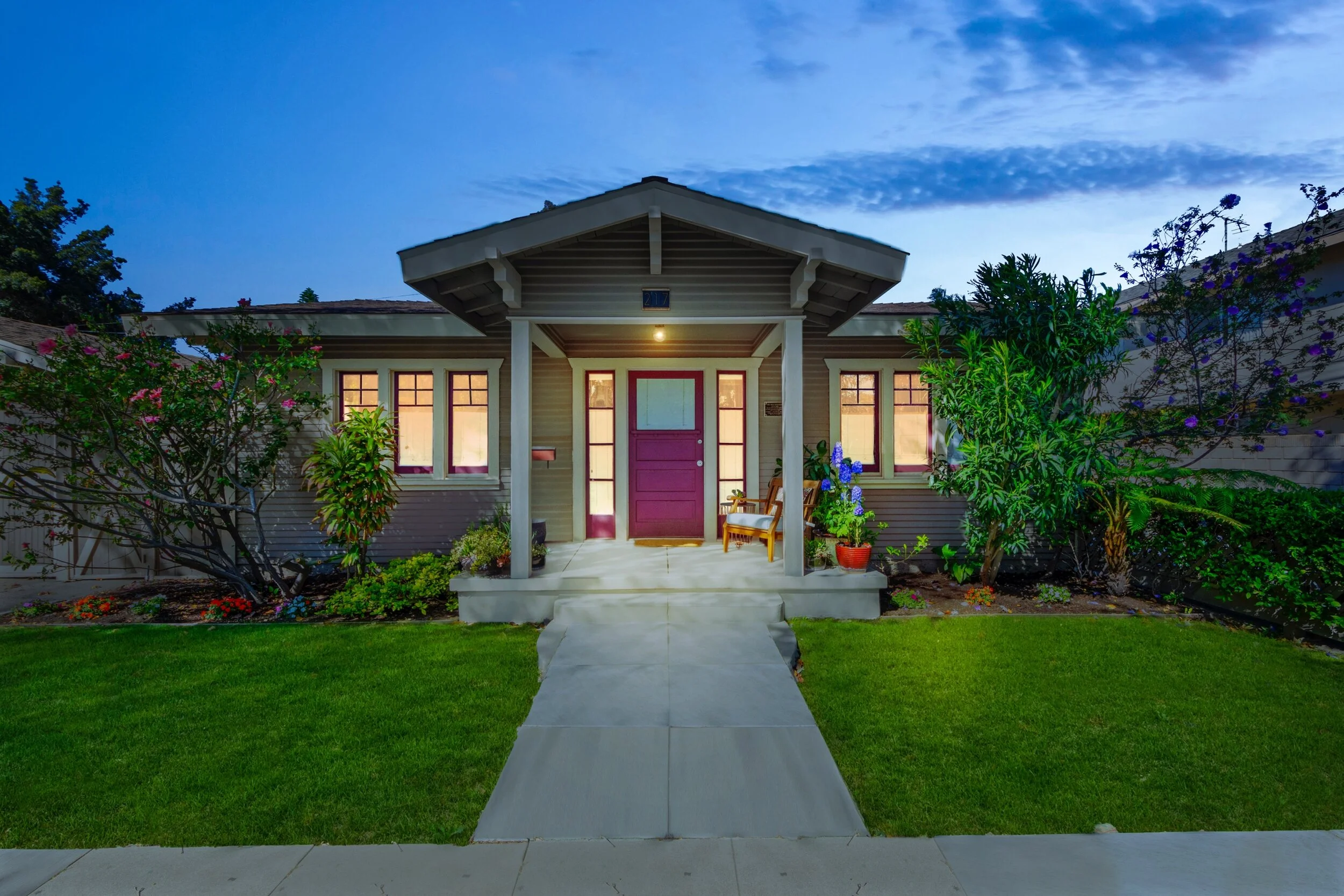 Front view of a cozy house with a purple front door, a small porch with a wooden chair, and lush green landscaping at dusk.