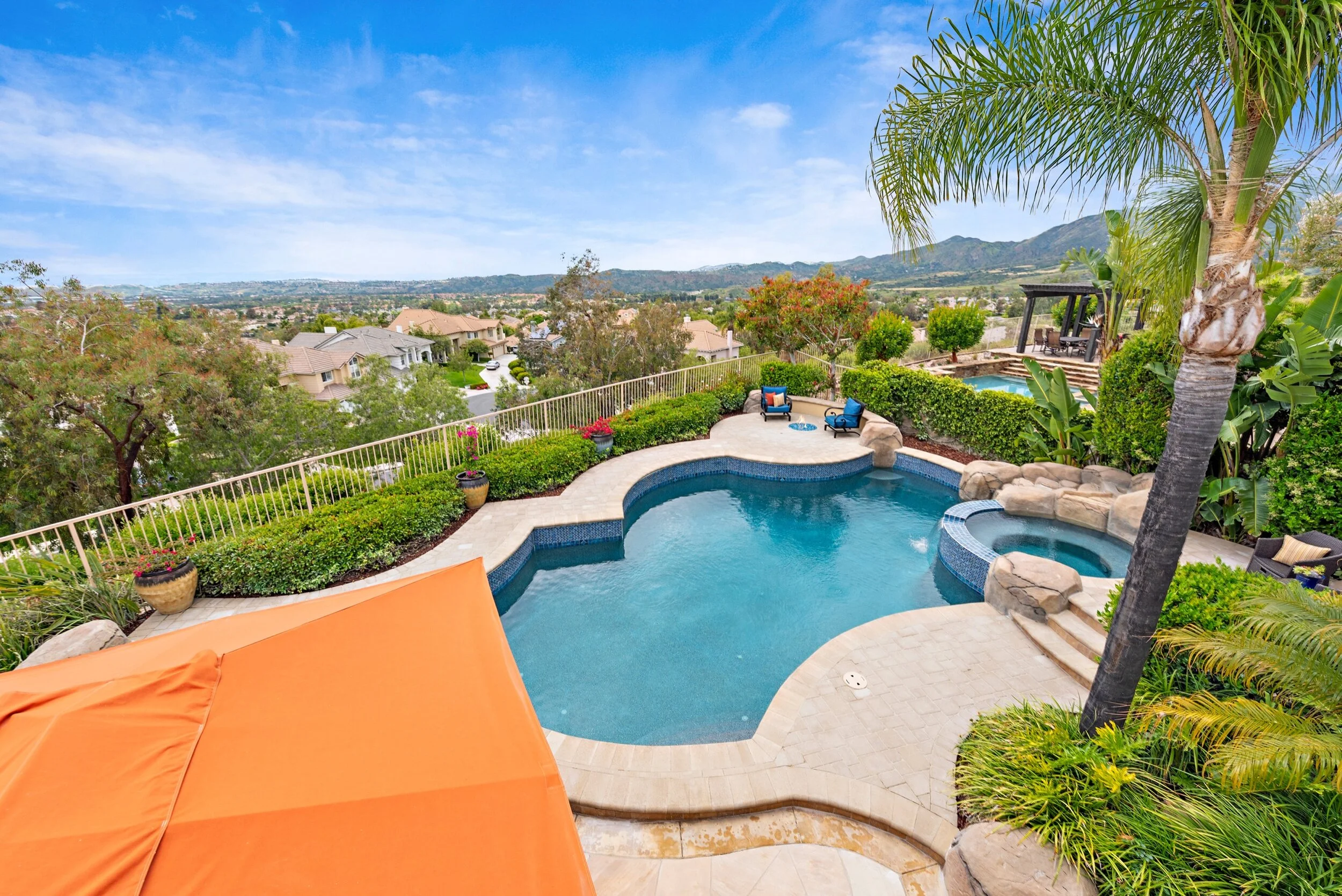 Backyard with swimming pool, surrounding lush greenery, potted plants, outdoor seating, and a scenic view of distant hills under a blue sky.