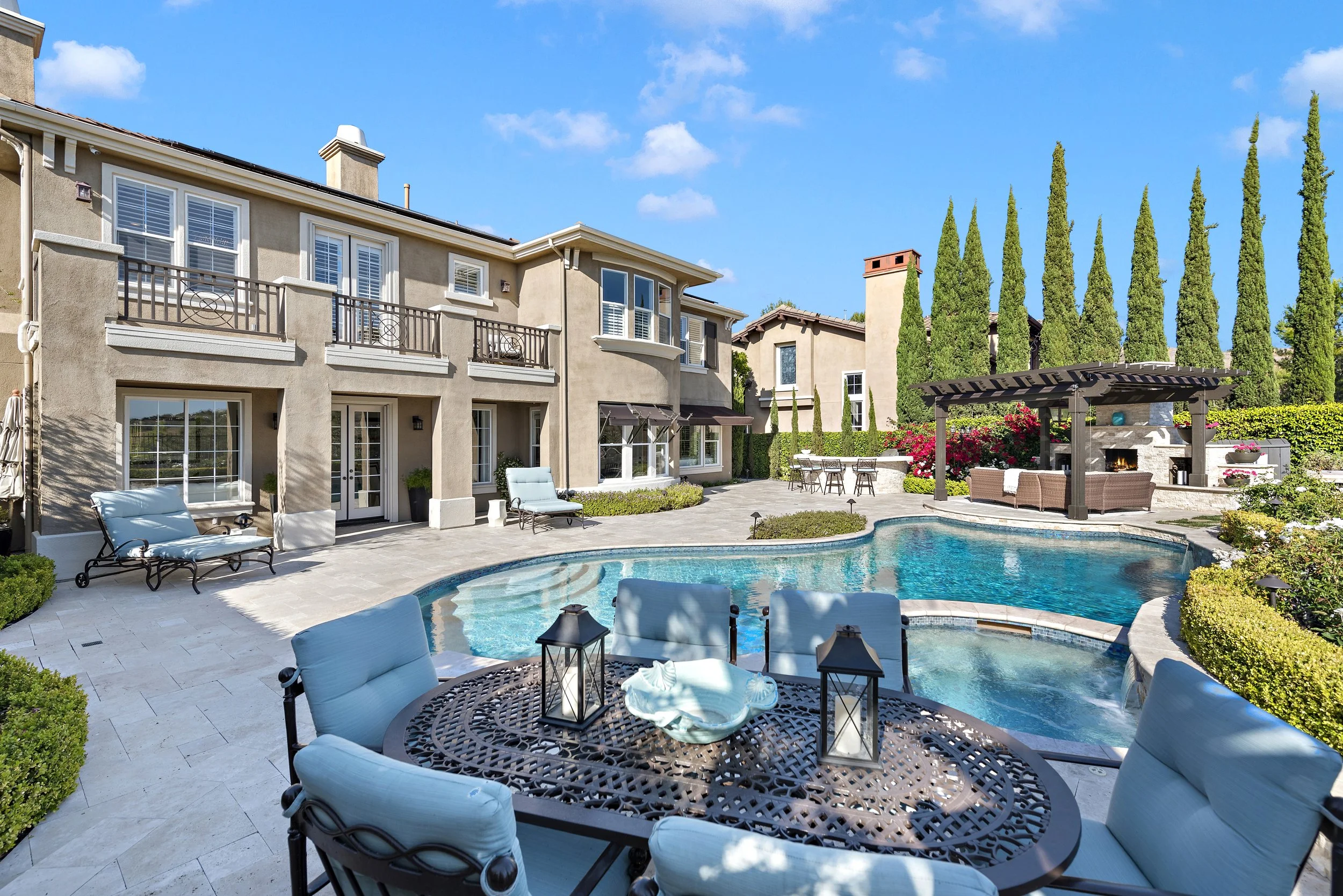 Backyard of a house with a swimming pool, patio furniture, and a fire pit with sitting area, surrounded by tall cypress trees and landscaped garden.