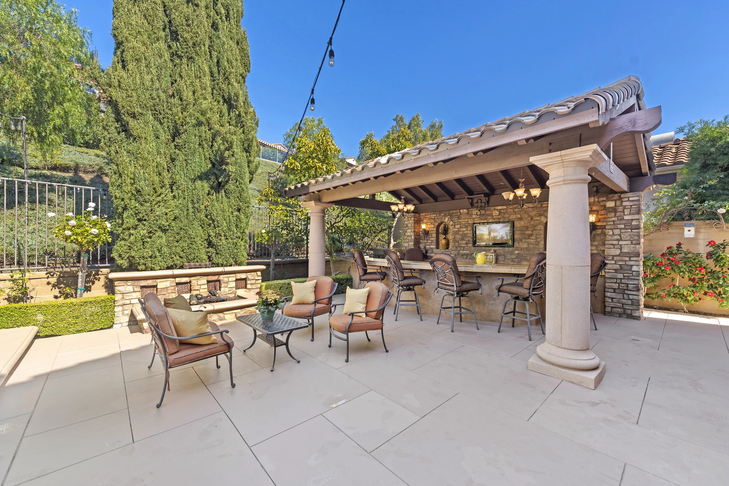 Outdoor patio with seating area, bar, and string lights under a blue sky, surrounded by greenery and privacy fencing.