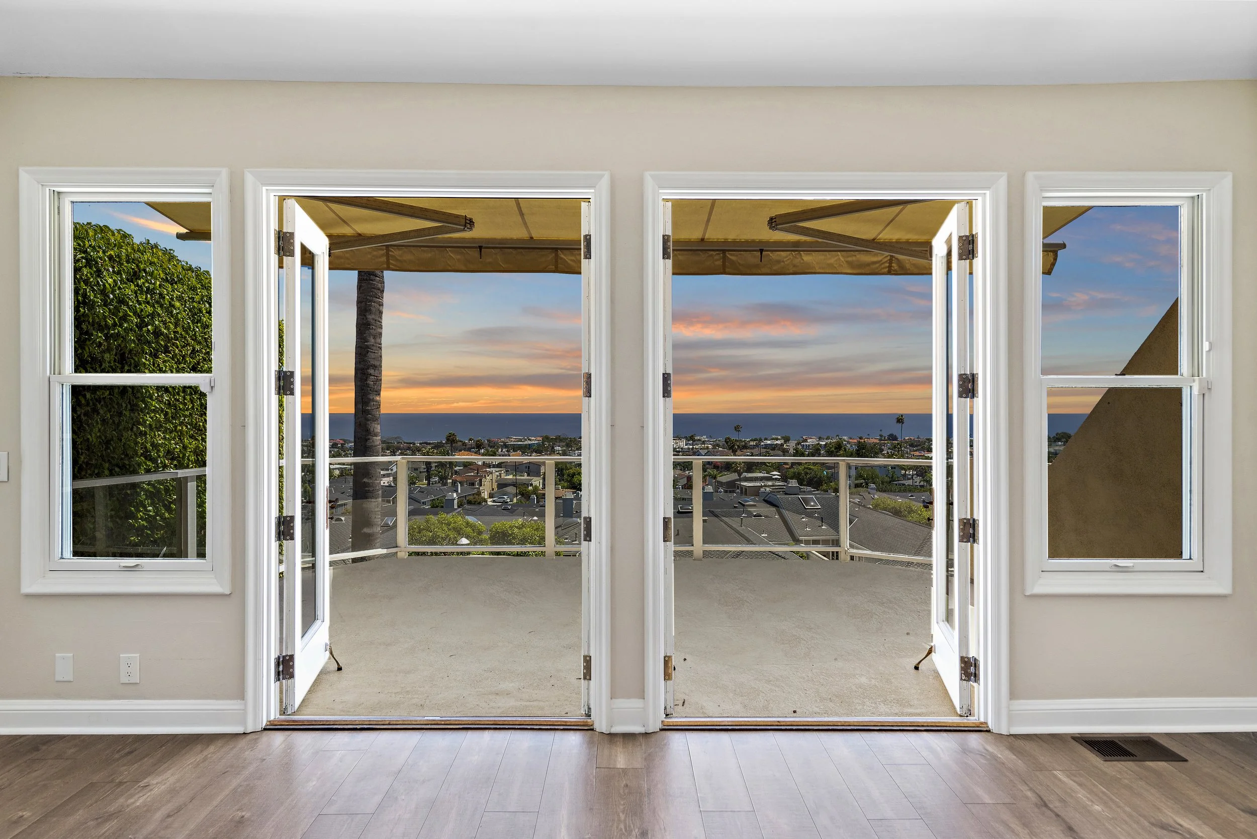 View through open double glass doors to a balcony at sunset, with rooftops, palm trees, and the ocean in the distance.