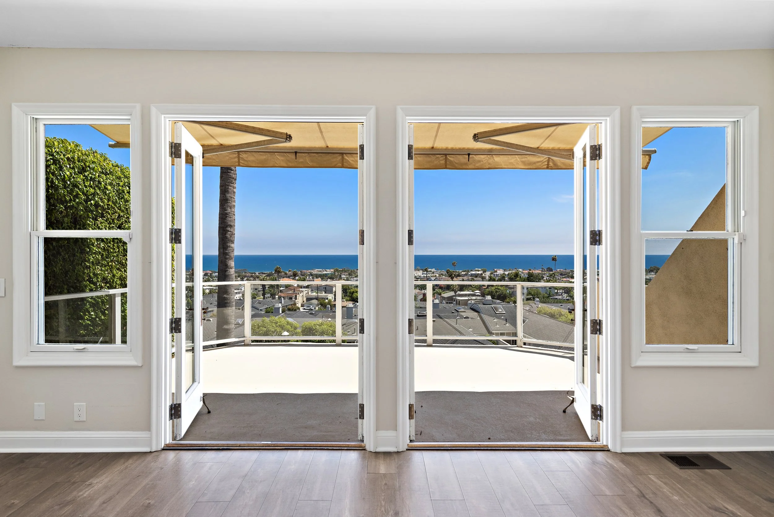 Room with open double doors leading to a balcony overlooking a cityscape with cars, trees, and the ocean in the distance under a blue sky.