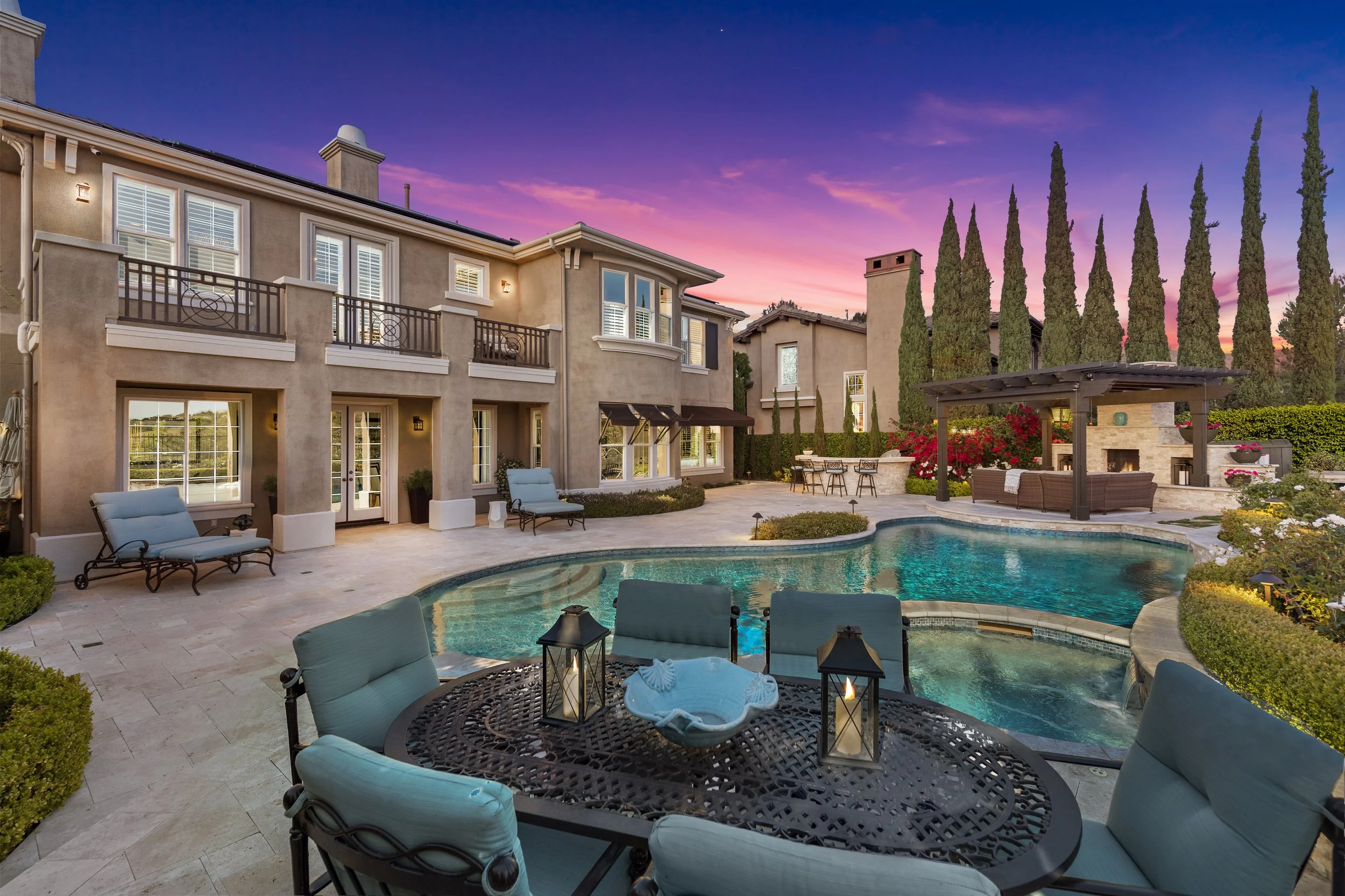 A backyard with a swimming pool, lounge chairs, a covered outdoor seating area, and a house with balconies is shown at sunset, with a sky painted in pink and purple hues and tall trees in the background.