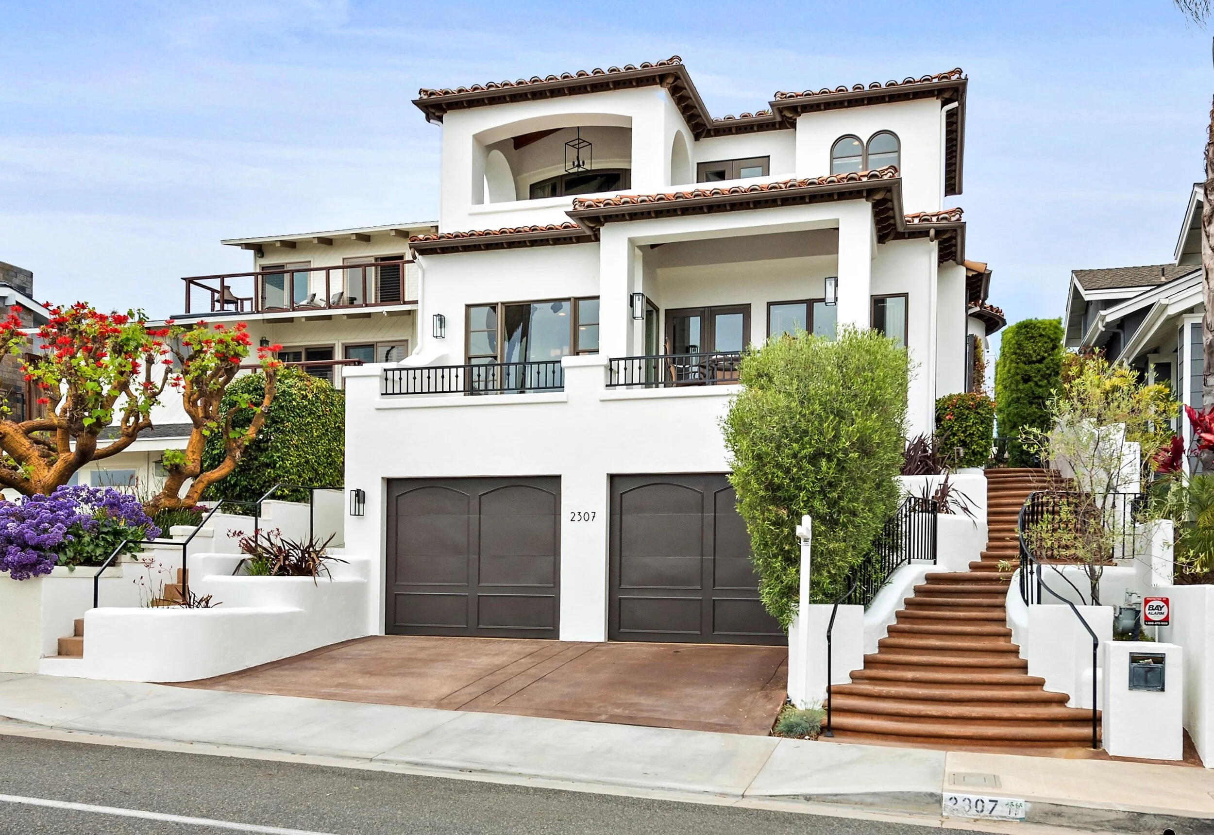 Modern multi-story white house with brown tiled roofs, surrounded by greenery and flowers, featuring multiple balconies, large windows, and a garage with two doors.
