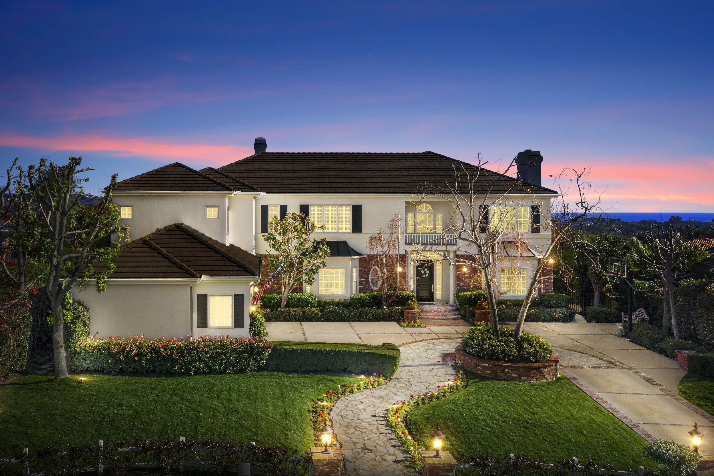 A large, two-story house with a well-lit front yard during twilight, featuring a curved stone pathway, manicured lawn, leafless trees, and outdoor lighting.