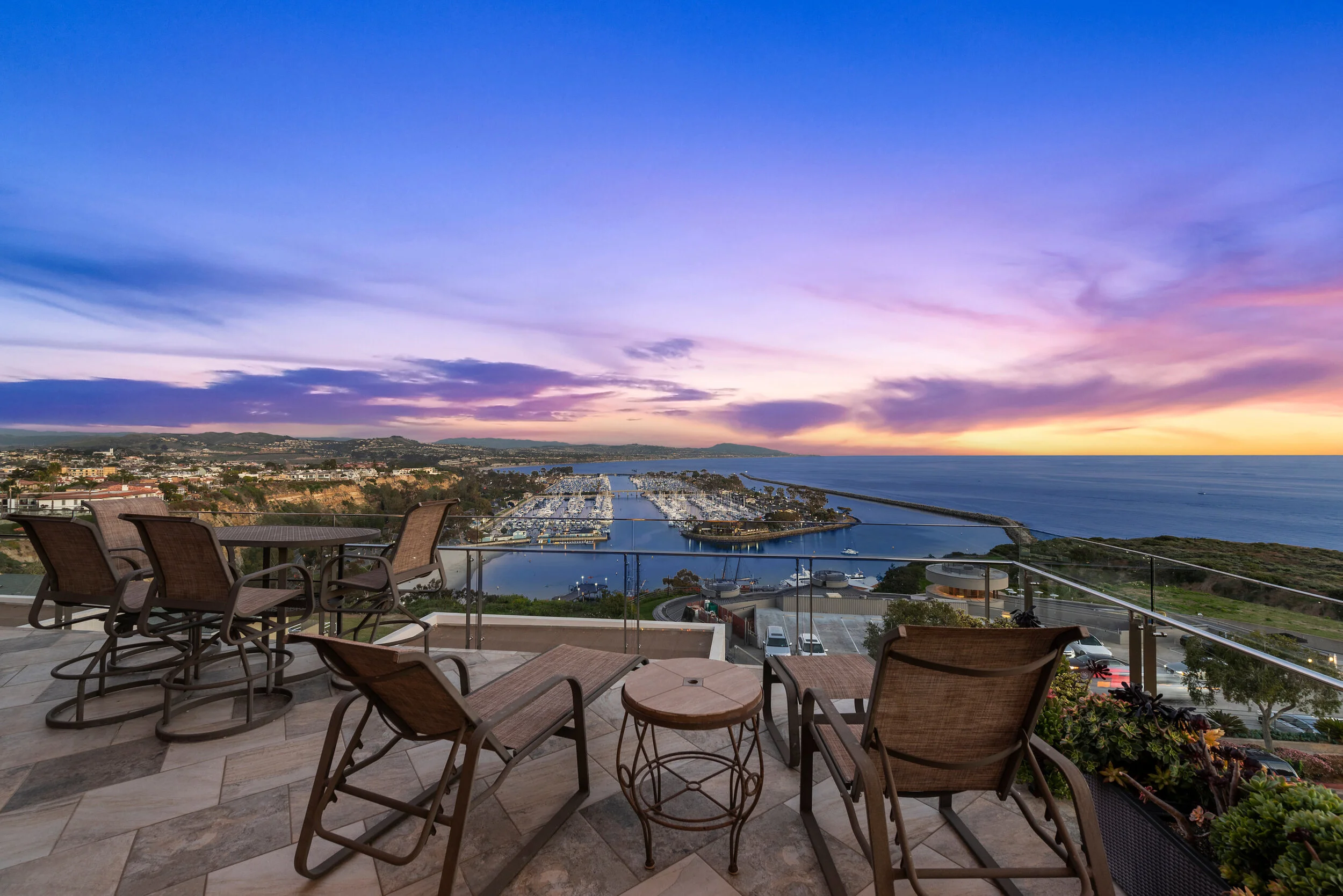 An outdoor balcony overlooking a marina at sunset, with chairs and a small table, and a cityscape in the distance.