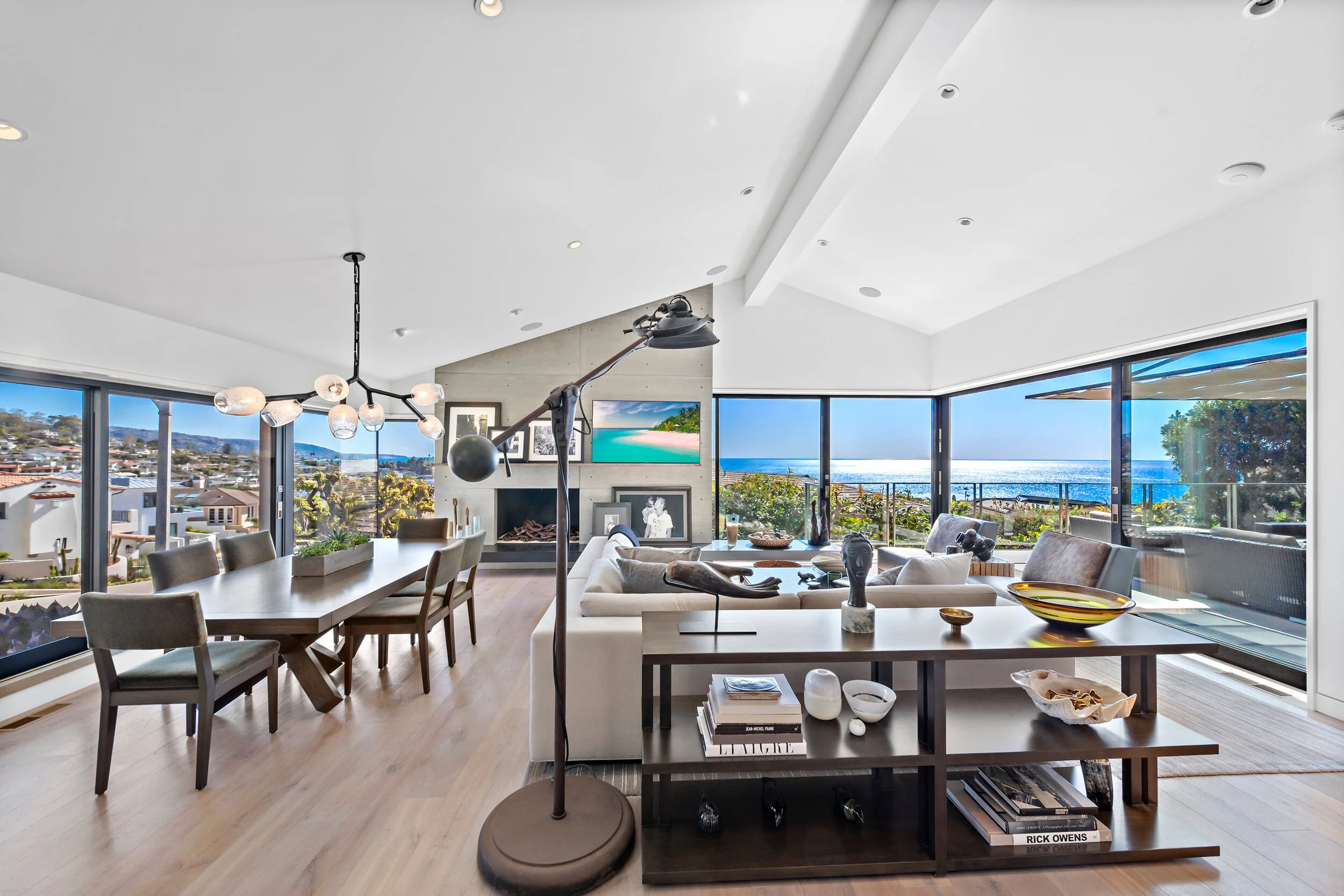 Open-concept living and dining room with large windows showing ocean view, modern furniture, fireplace, and decorative art pieces.