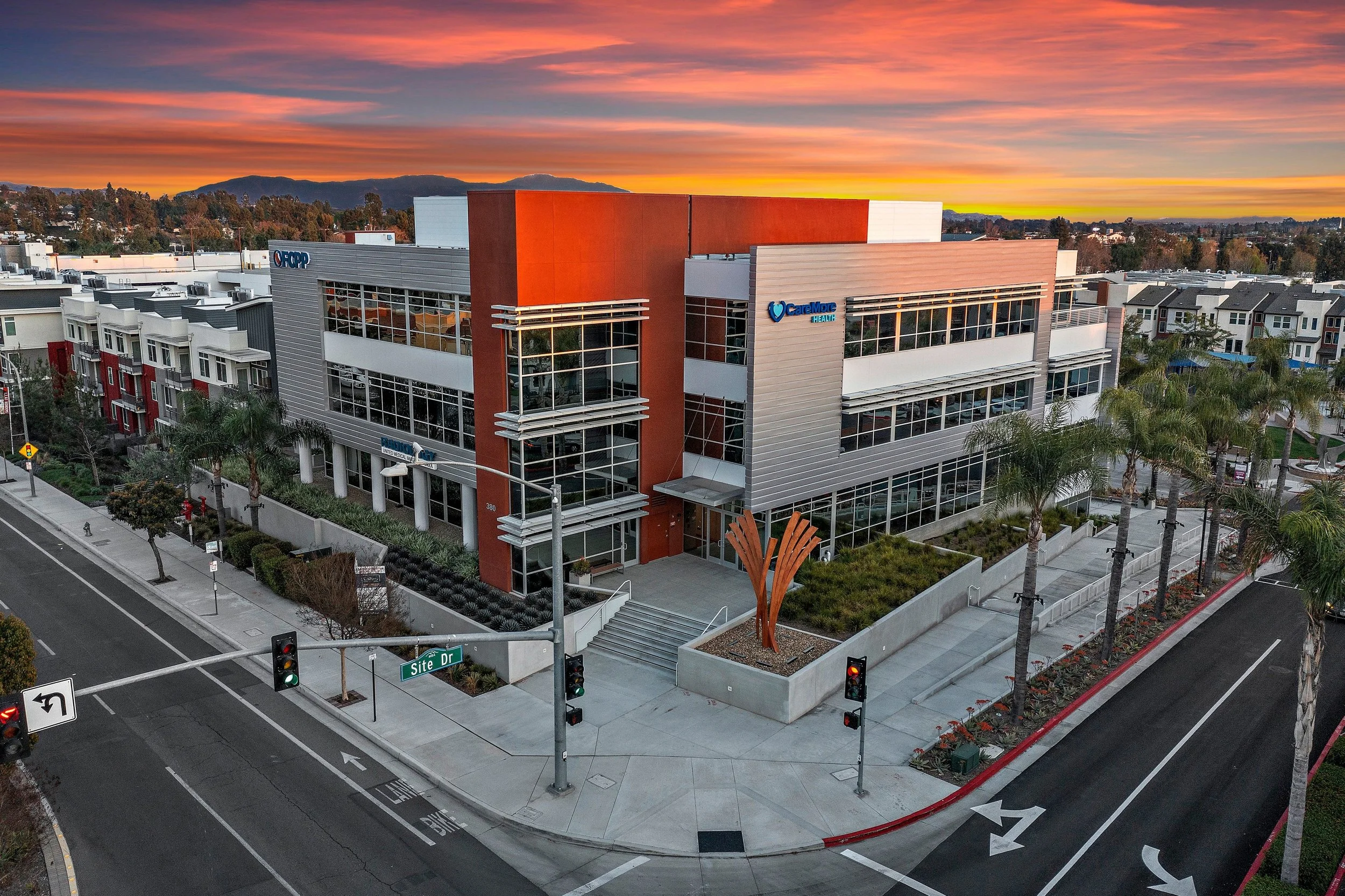 A modern multi-story healthcare building at sunset with palm trees in the foreground and a colorful sky in the background.