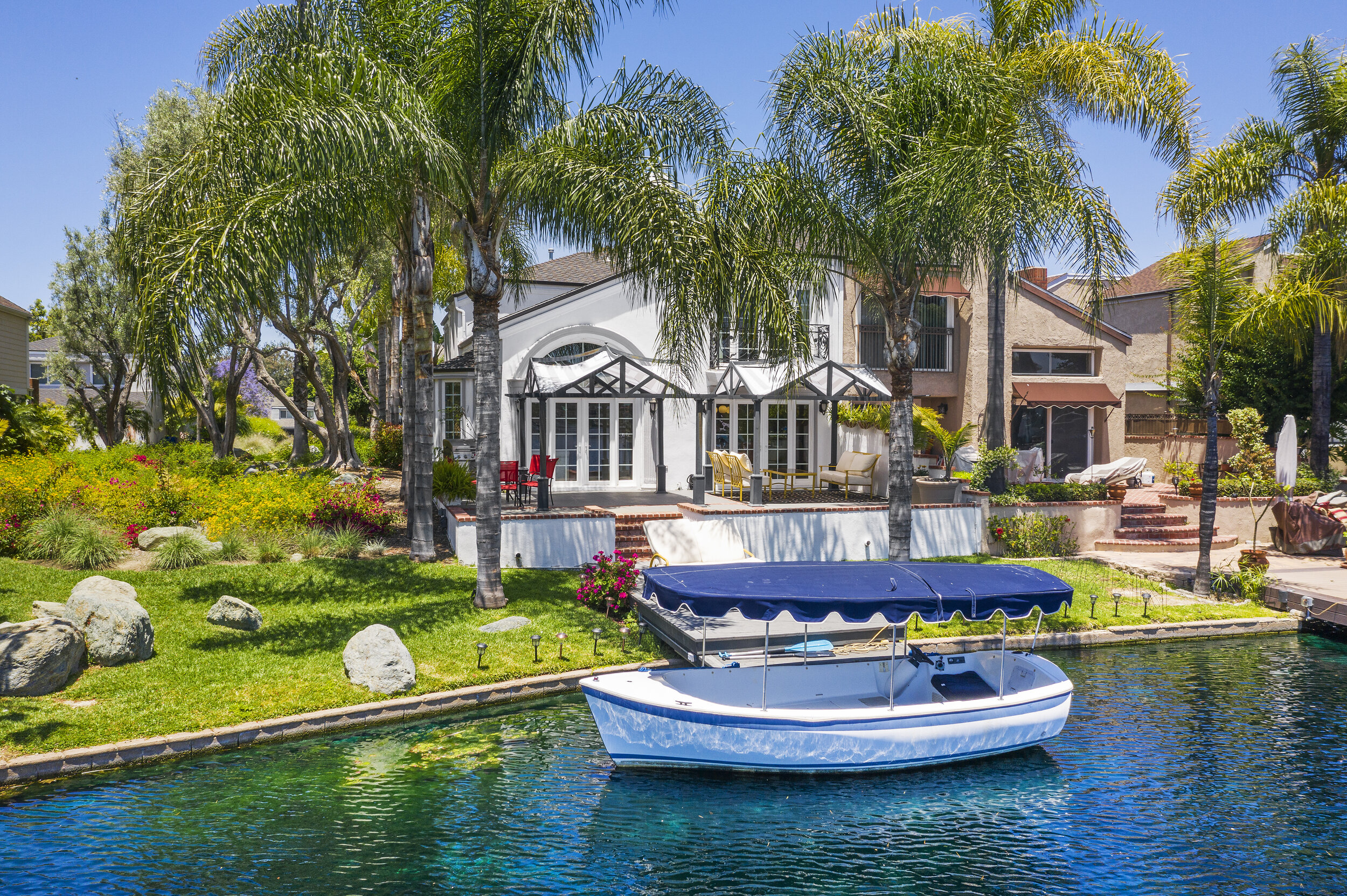 A waterfront backyard with a boat docked in the water, close to the lush green lawn with large rocks, tall palm trees, colorful flower beds, and a multi-story house with patio furniture and umbrellas.