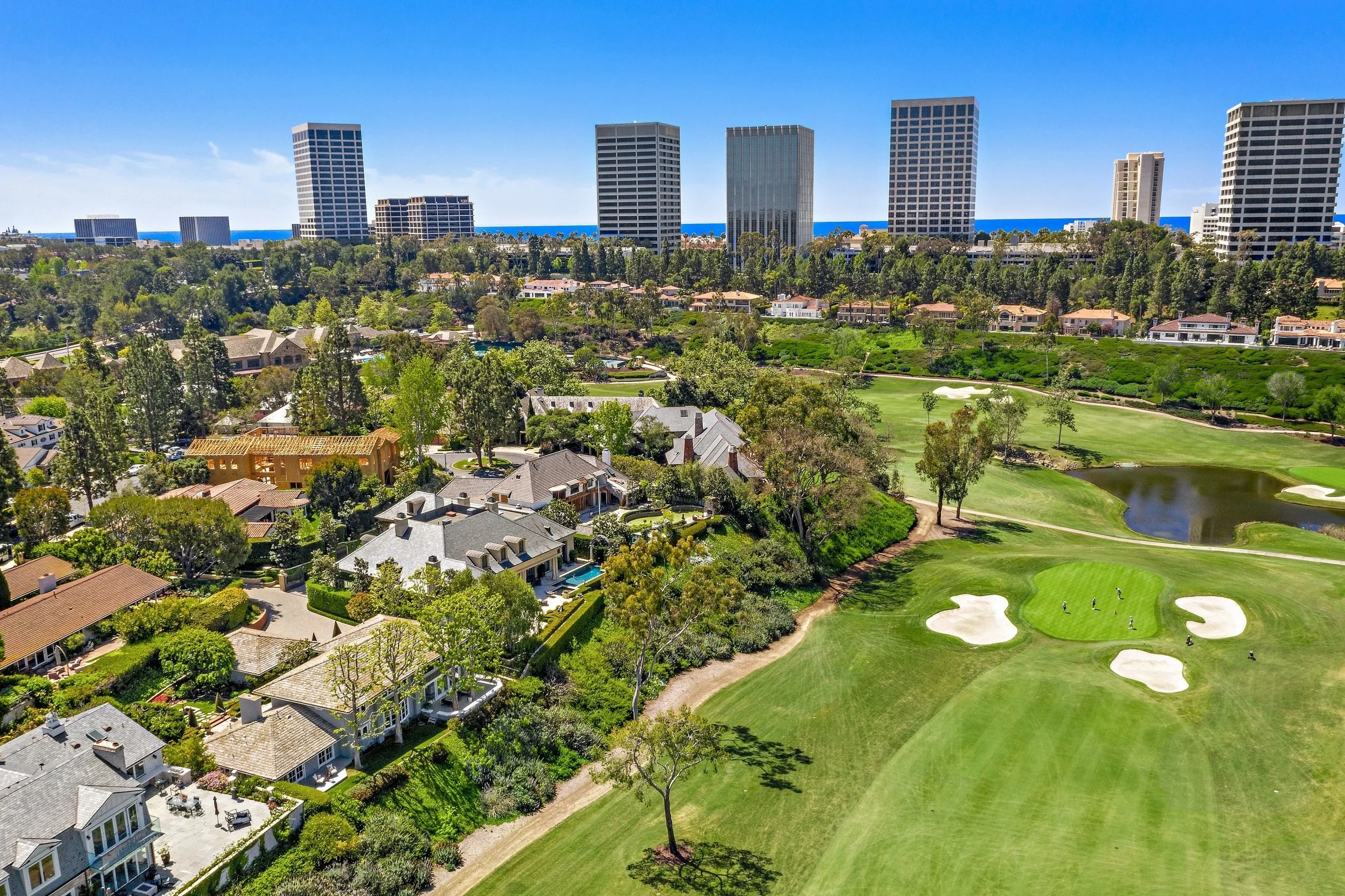 Aerial view of a golf course with sand bunkers and water hazards, surrounded by residential houses with a cityscape of tall buildings in the background.