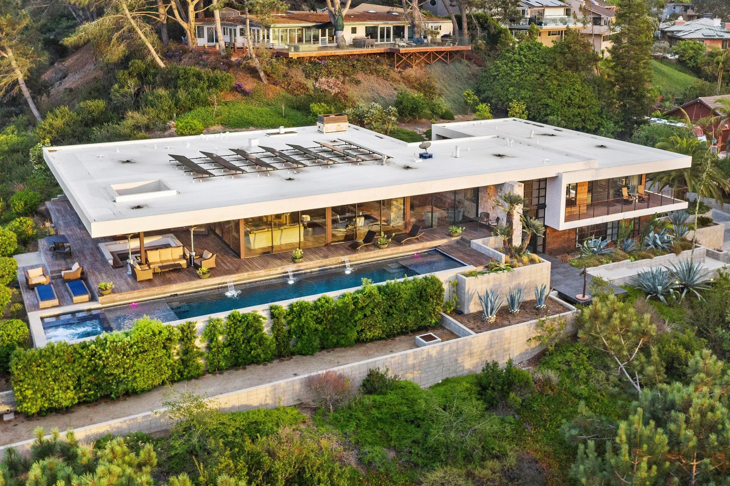 A modern hillside house with a flat roof, large glass walls, an outdoor pool, and surrounding lush greenery.