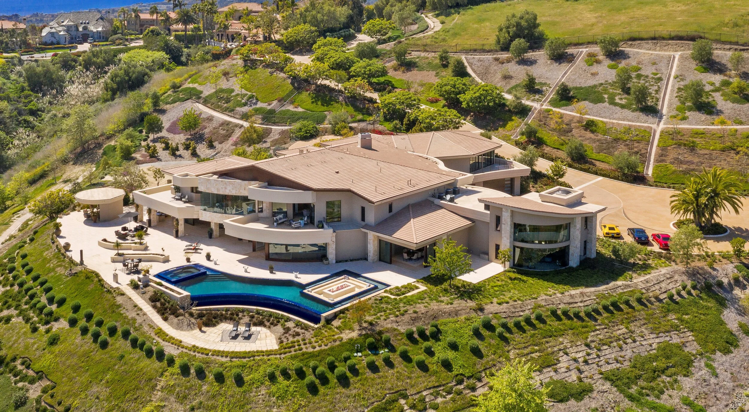 Aerial view of a large modern house with a swimming pool, hot tub, outdoor seating areas, and a landscaped yard on a hillside with winding paths and neighboring homes in the distance.