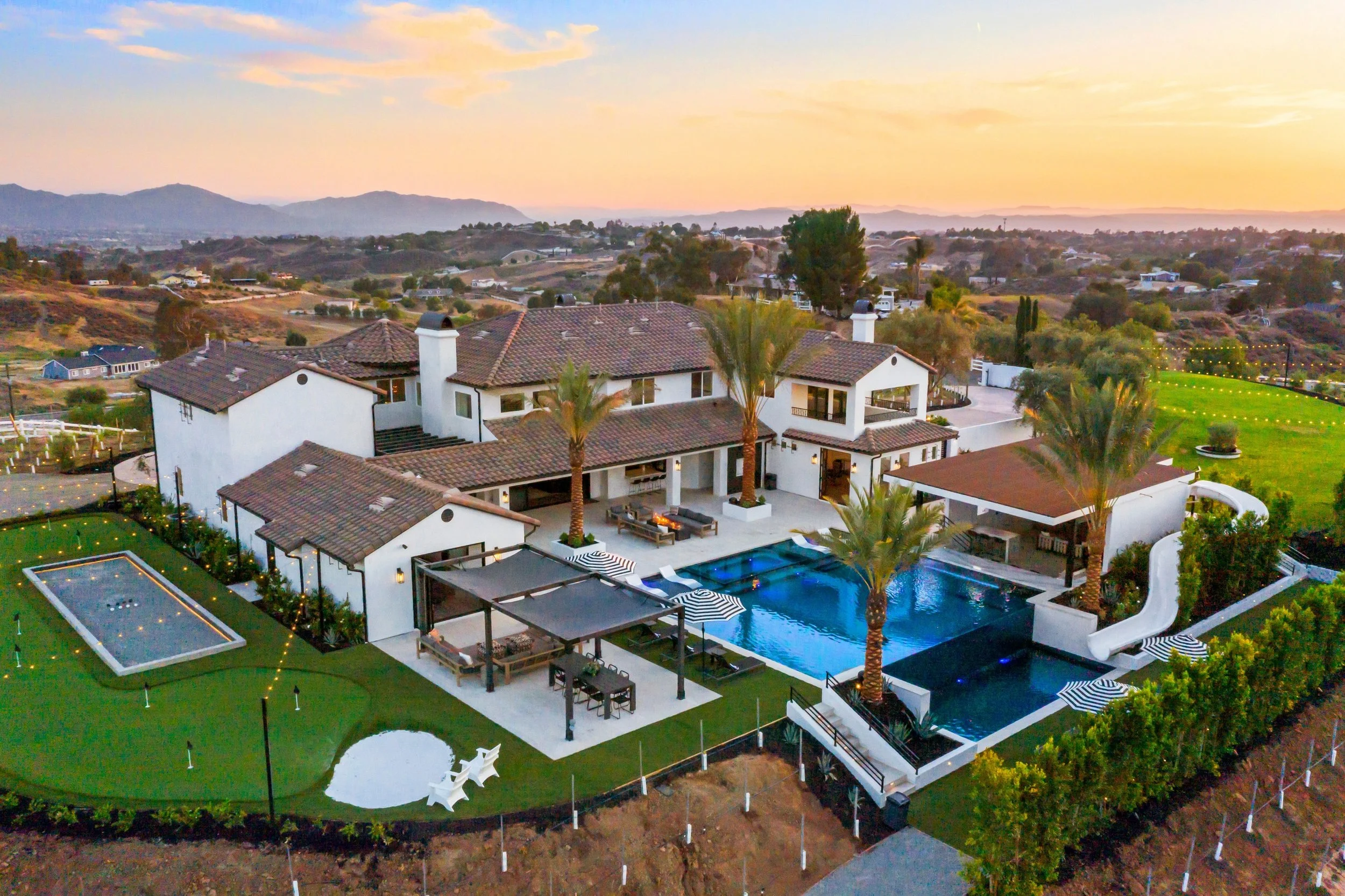 A large, luxurious house with multiple levels, surrounded by a well-maintained yard, tall palm trees, and a swimming pool area featuring a slide and loungers, set against a sunset sky in a hilly landscape.