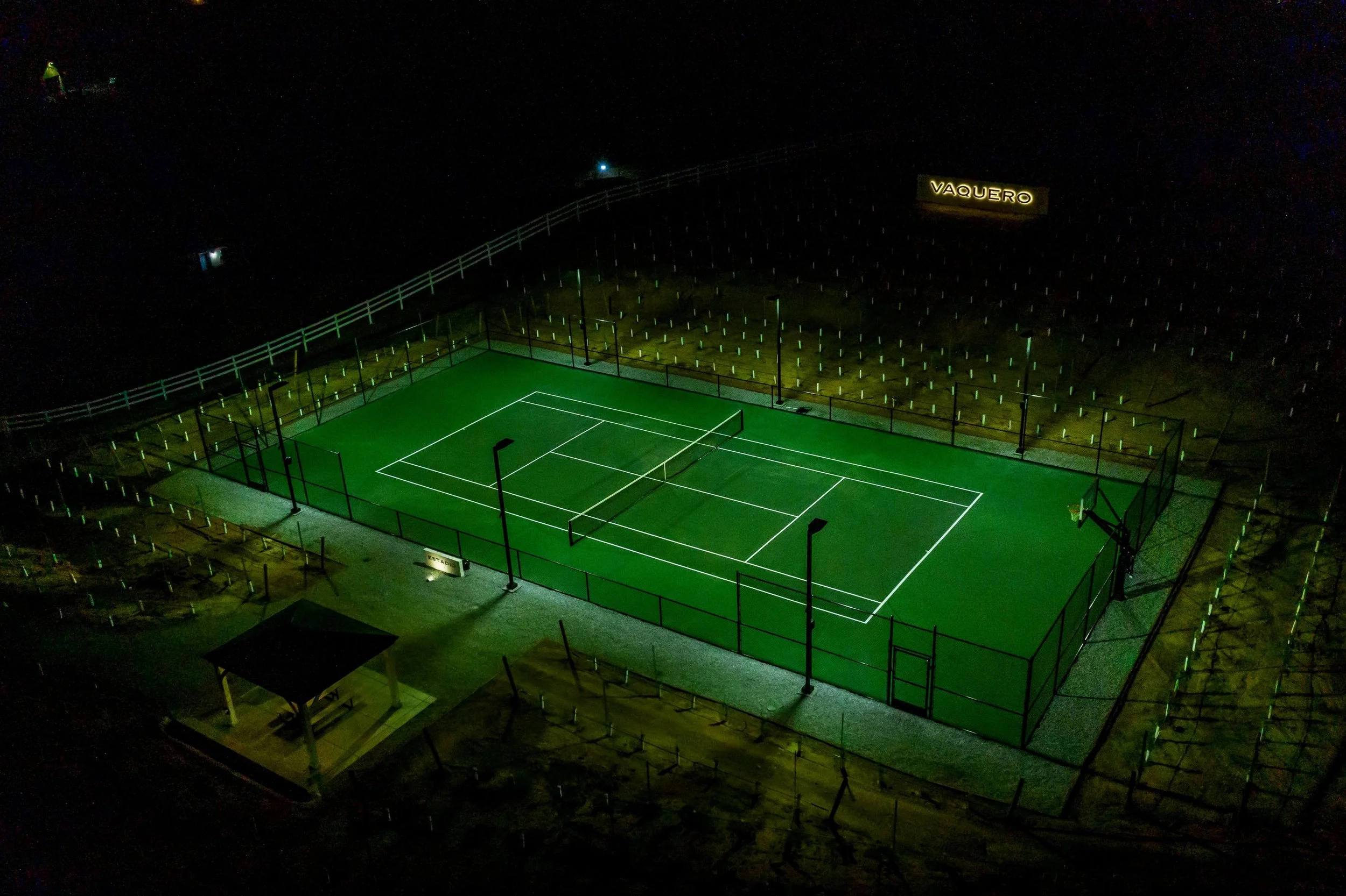 A brightly lit outdoor tennis court at night, surrounded by a tall fence and seating area, with a sign reading 'Vaquero' in the background.