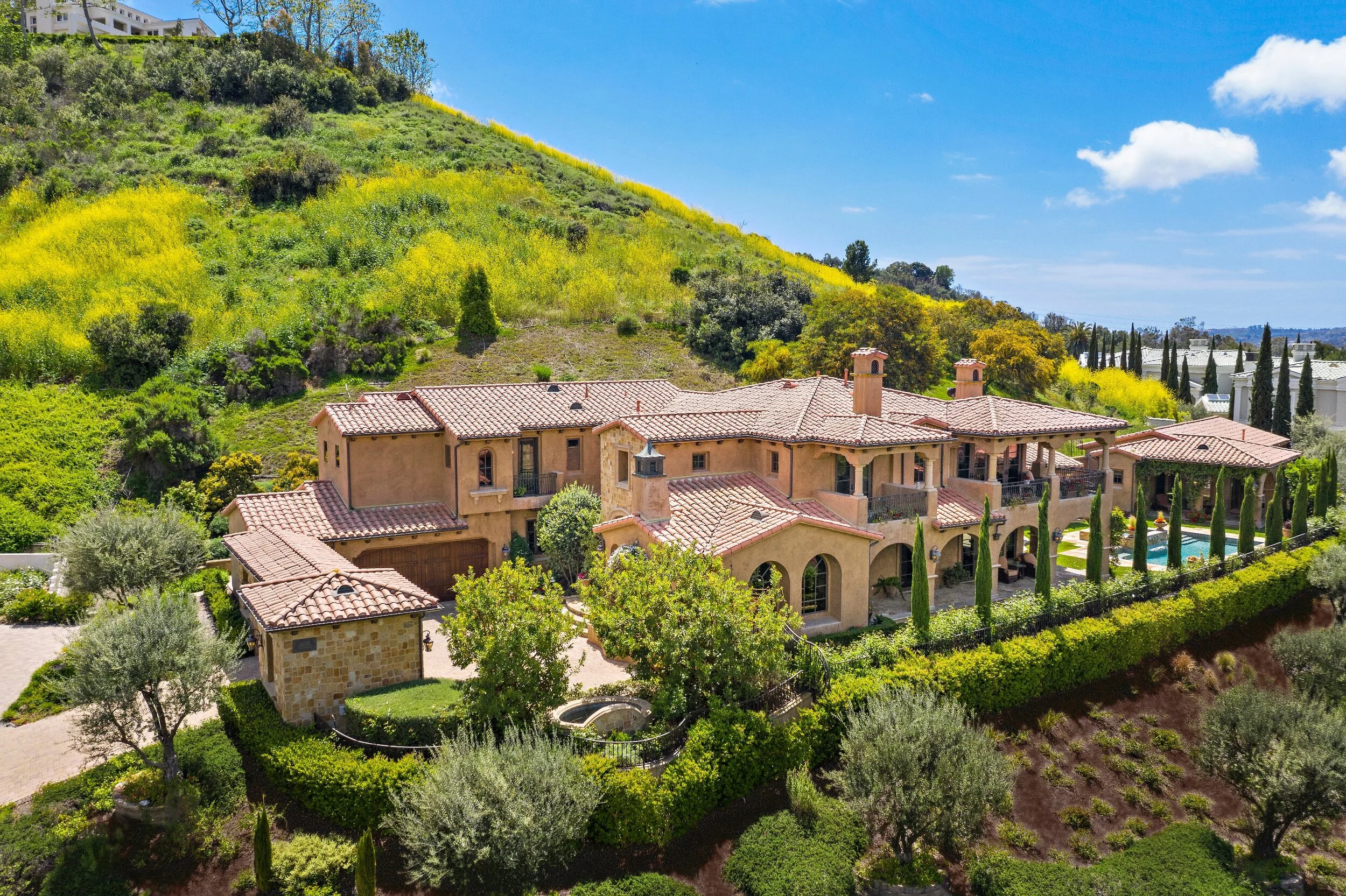 Large Mediterranean-style house with red tile roof, lush green trees, and landscaped yard on hillside under blue sky.