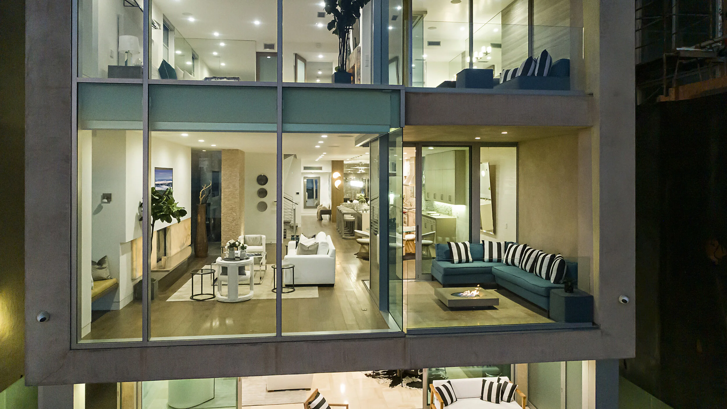 Interior view of a modern multi-story house with large glass windows, showcasing a living room with white sofas, a fireplace, blue sectional sofa with striped pillows, and a dining area with a kitchen in the background.