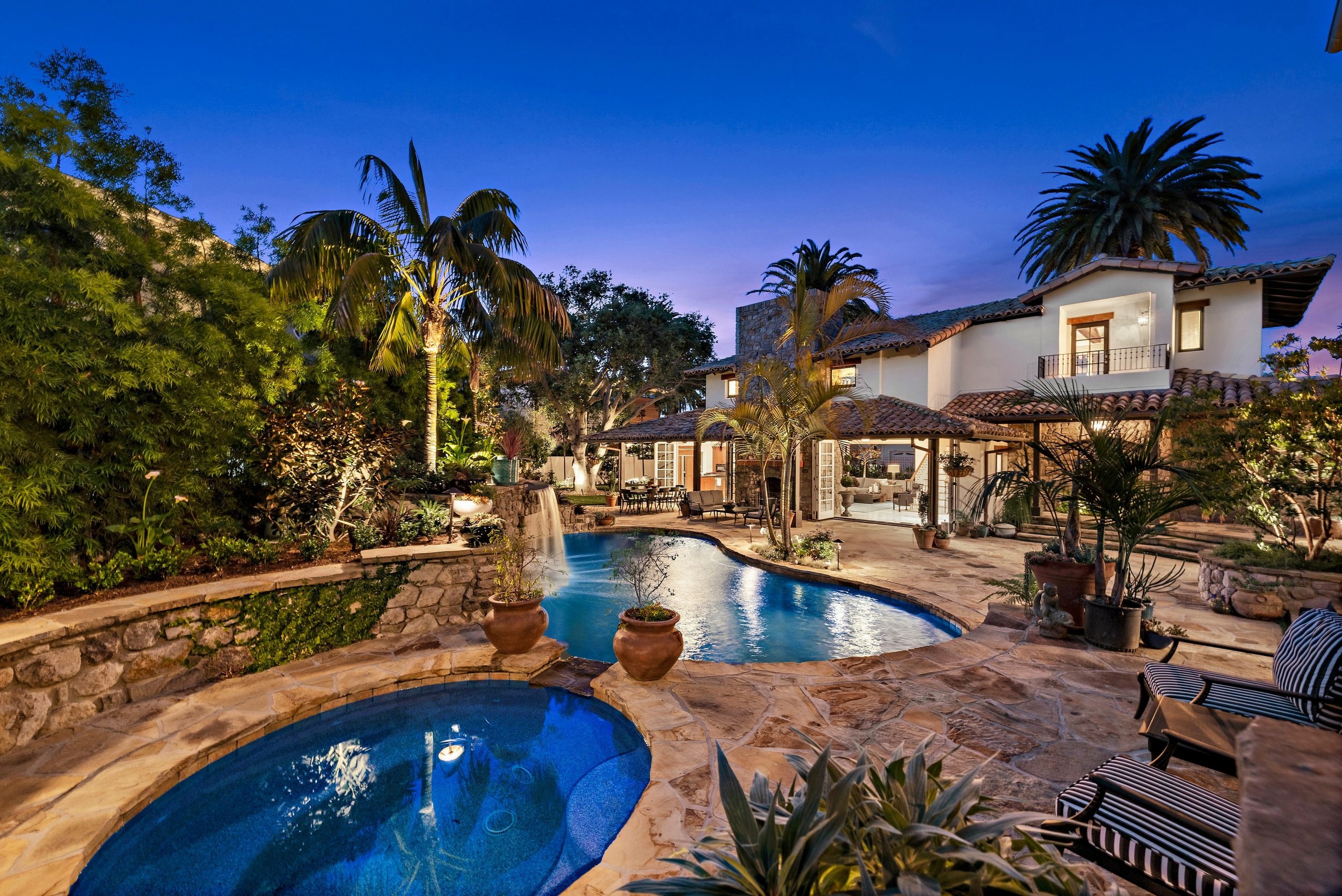Luxury backyard with swimming pool, hot tub, patio, lush palm trees, and a two-story house with Spanish tile roof during dusk.