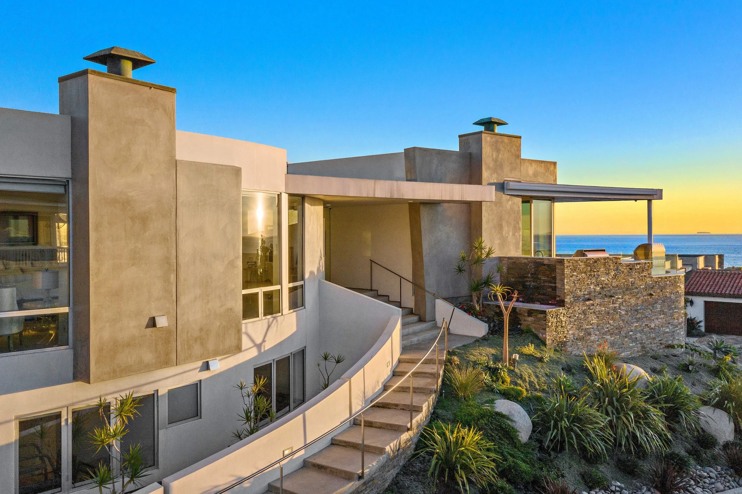 Modern house with outdoor staircase leading to the entrance, overlooking the ocean at sunset, with landscaped garden and clear sky.