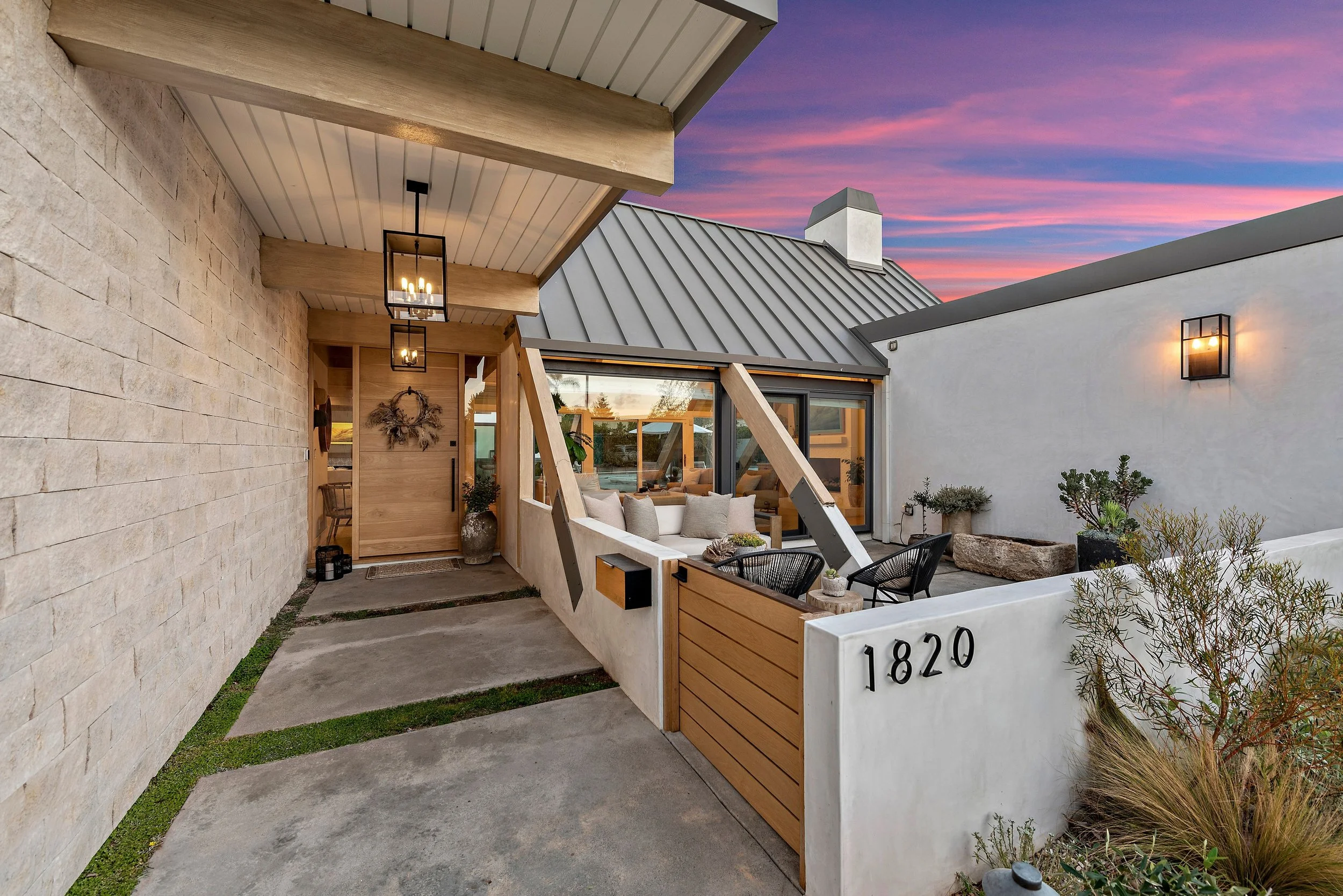 Modern house entry with a small patio, featuring a mix of stone, wood, and white walls, with large glass windows, potted plants, outdoor seating, and a sunset sky in the background.