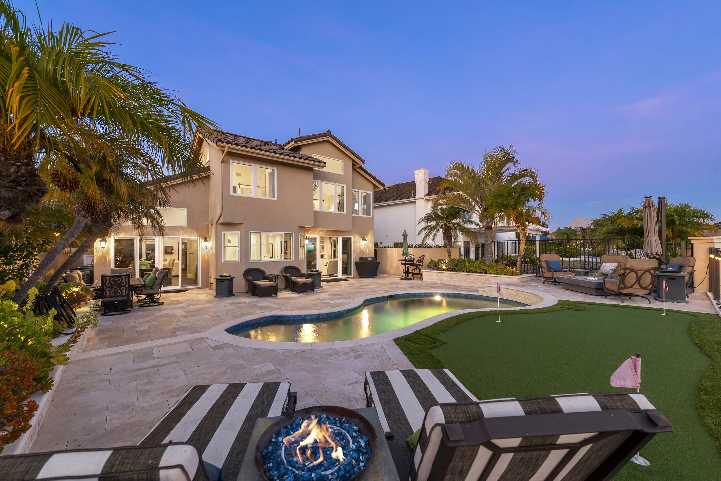 Backyard view of a modern house with a swimming pool, putting green, and outdoor furniture at dusk. Palm trees line the yard, and string lights illuminate the patio area.