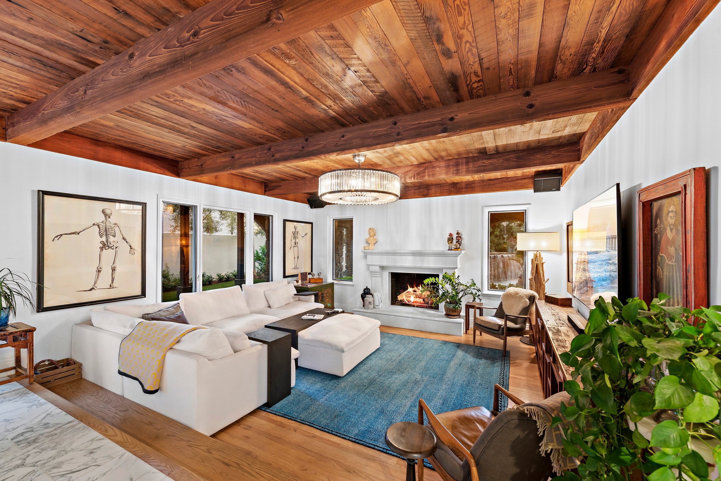 Living room with white sectional sofa, fireplace, art pieces, large blue rug, wooden ceiling beams, and large windows.