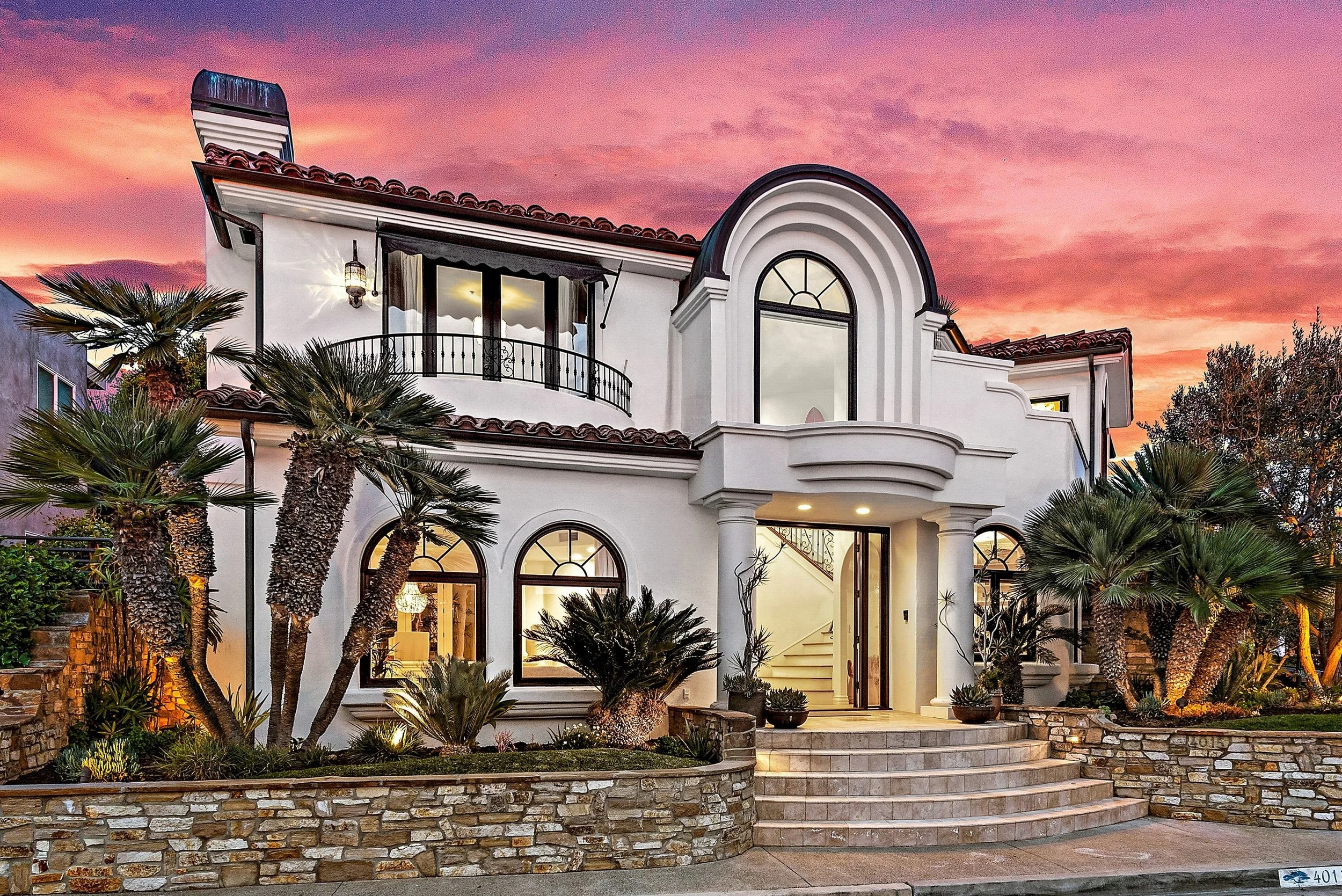 A luxurious white two-story house with arched windows, a rounded balcony, and tropical landscaping, viewed at sunset.