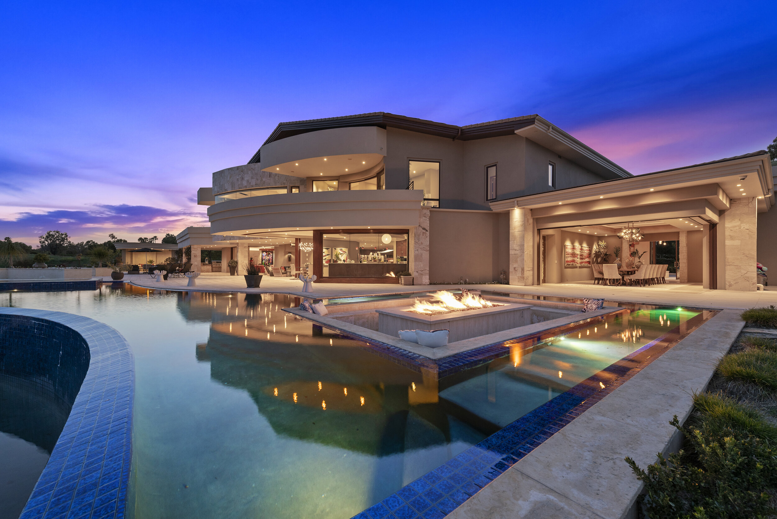 Luxury modern house with large swimming pool, fire feature, and outdoor dining area during sunset.