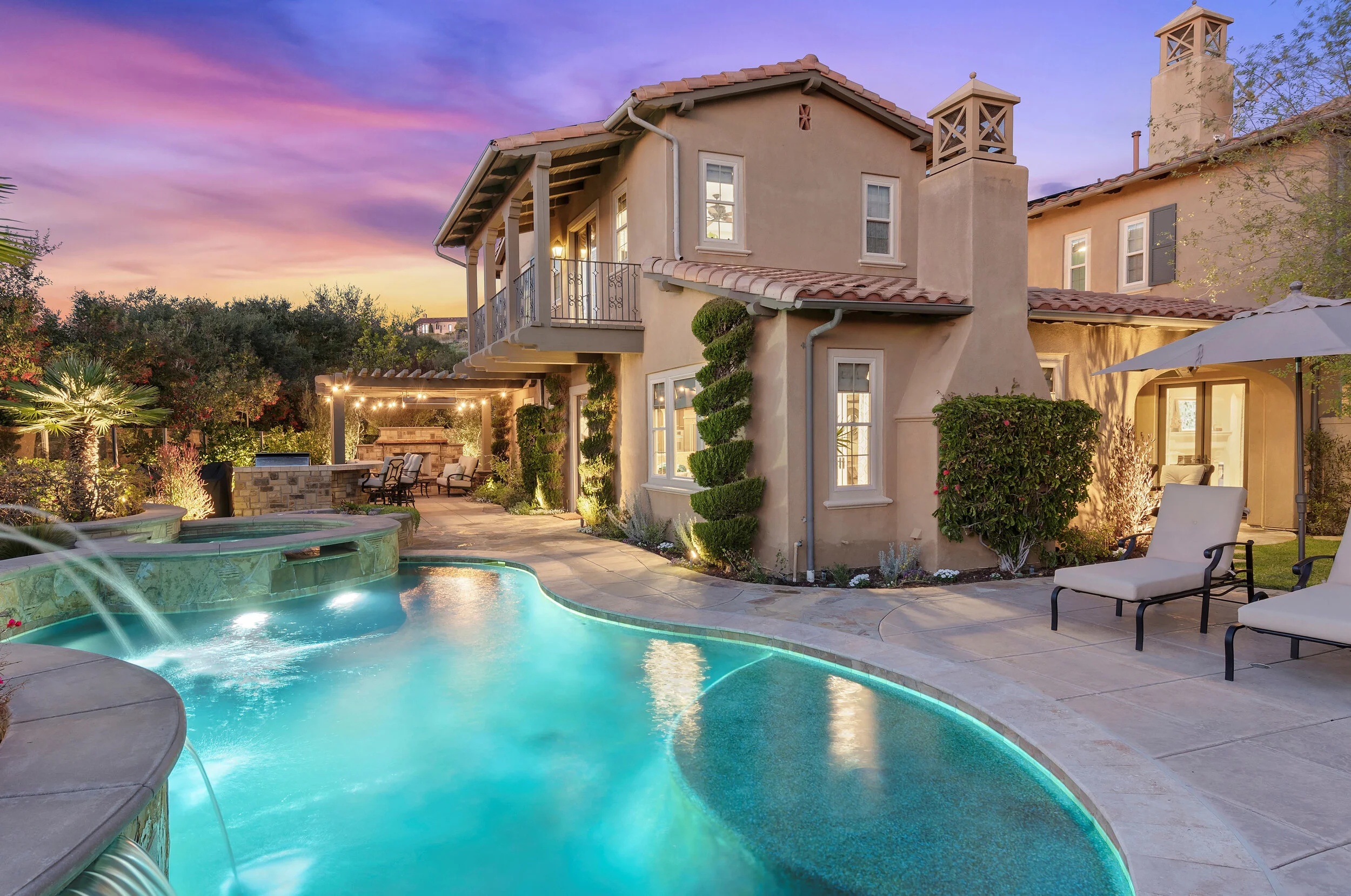 Luxury two-story house with a backyard pool area at sunset, featuring lounge chairs, a shaded outdoor seating area, and landscaped greenery.