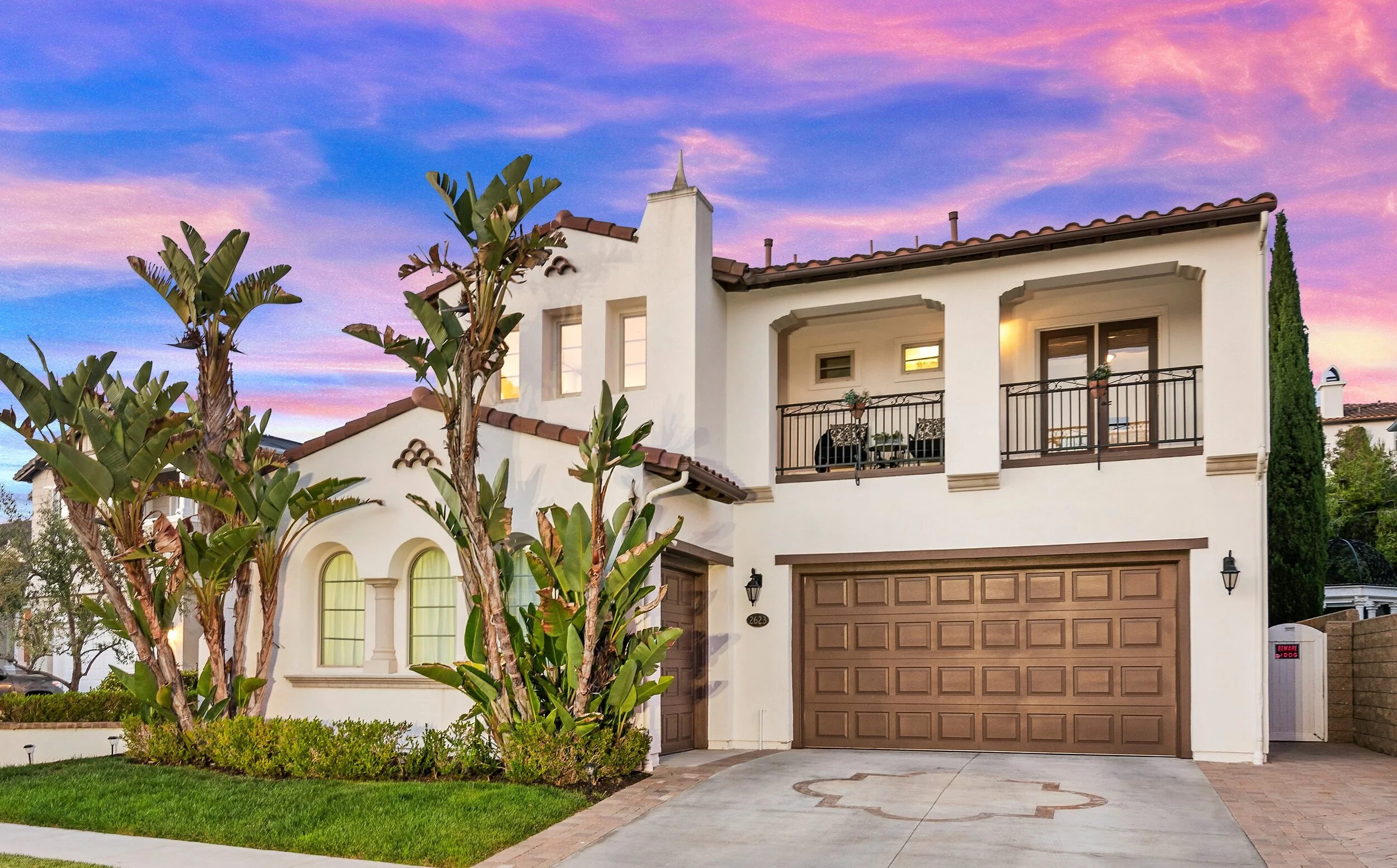 A two-story house with a white exterior, brown garage door, and a balcony with black railings, surrounded by green plants and trees under a colorful sunset sky.