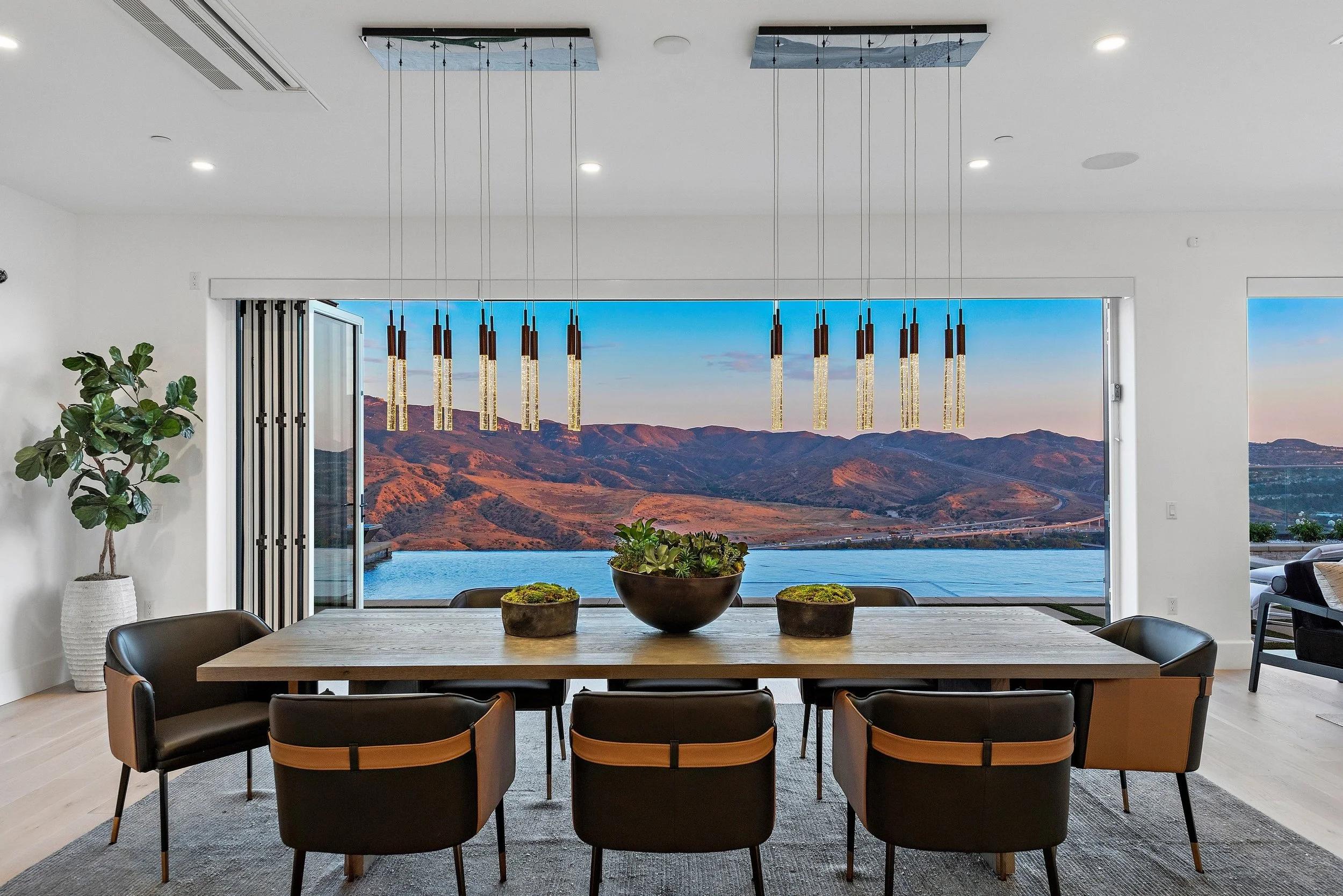 Modern dining room with a large wooden table, black chairs with tan accents, and a view of mountains and water through open sliding doors. Hanging pendant lights above the table and a potted plant in the corner.